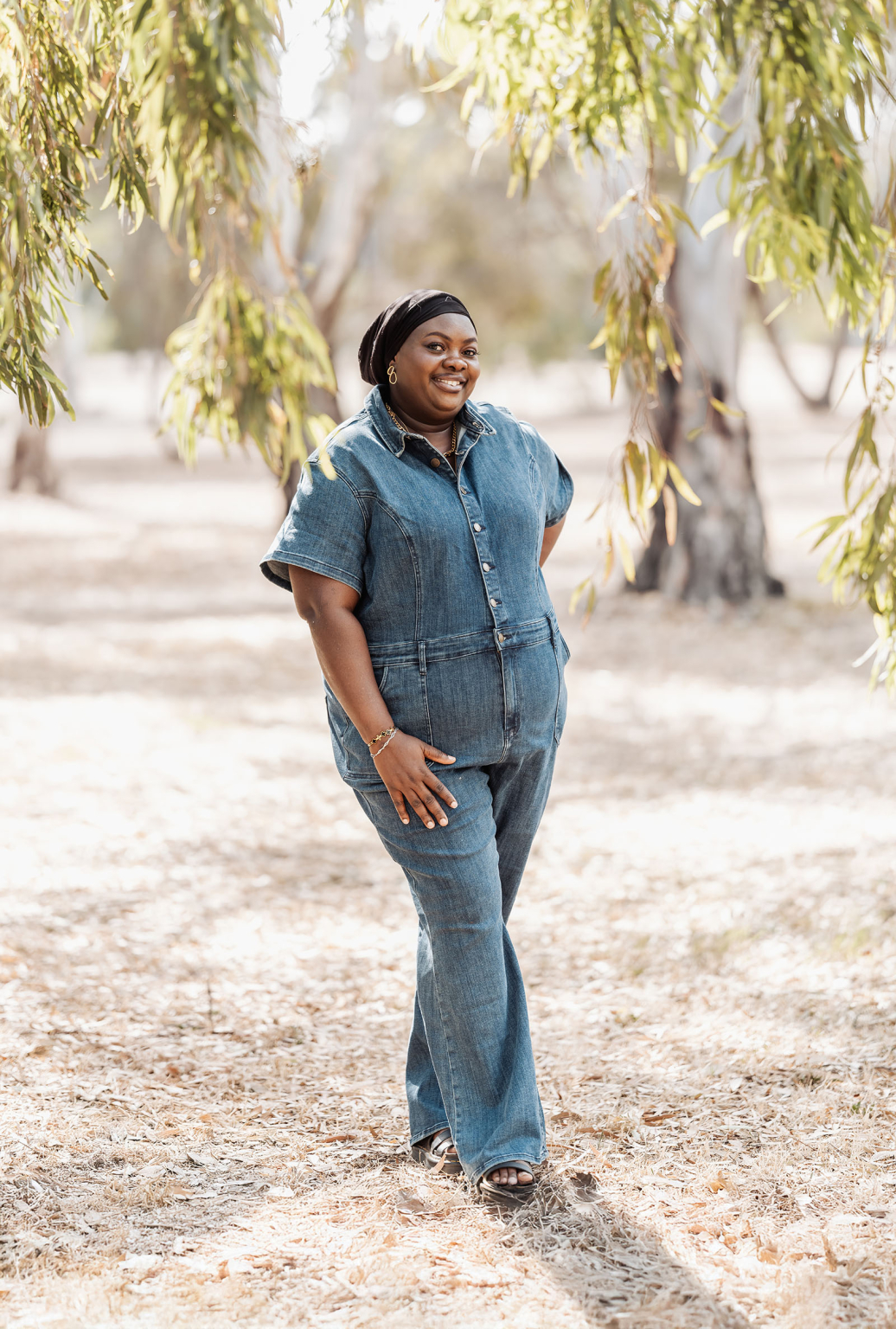 family photoshoot in veale gardens 25 Woman in denim jumpsuit smiling outdoors in a sunny, tree-filled park.