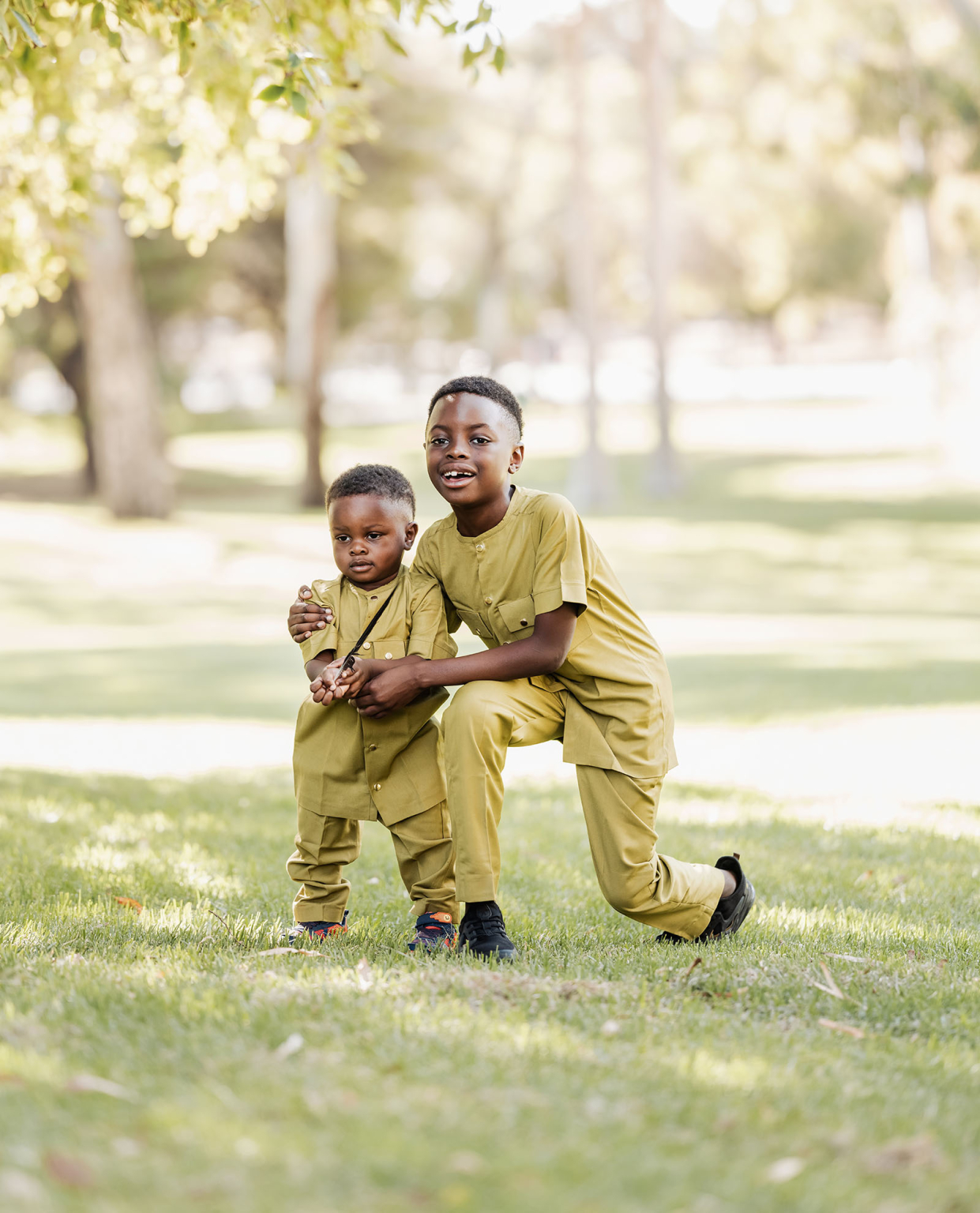 family photoshoot in veale gardens 19 Two children in matching outfits enjoy a sunny park, surrounded by trees and grass.