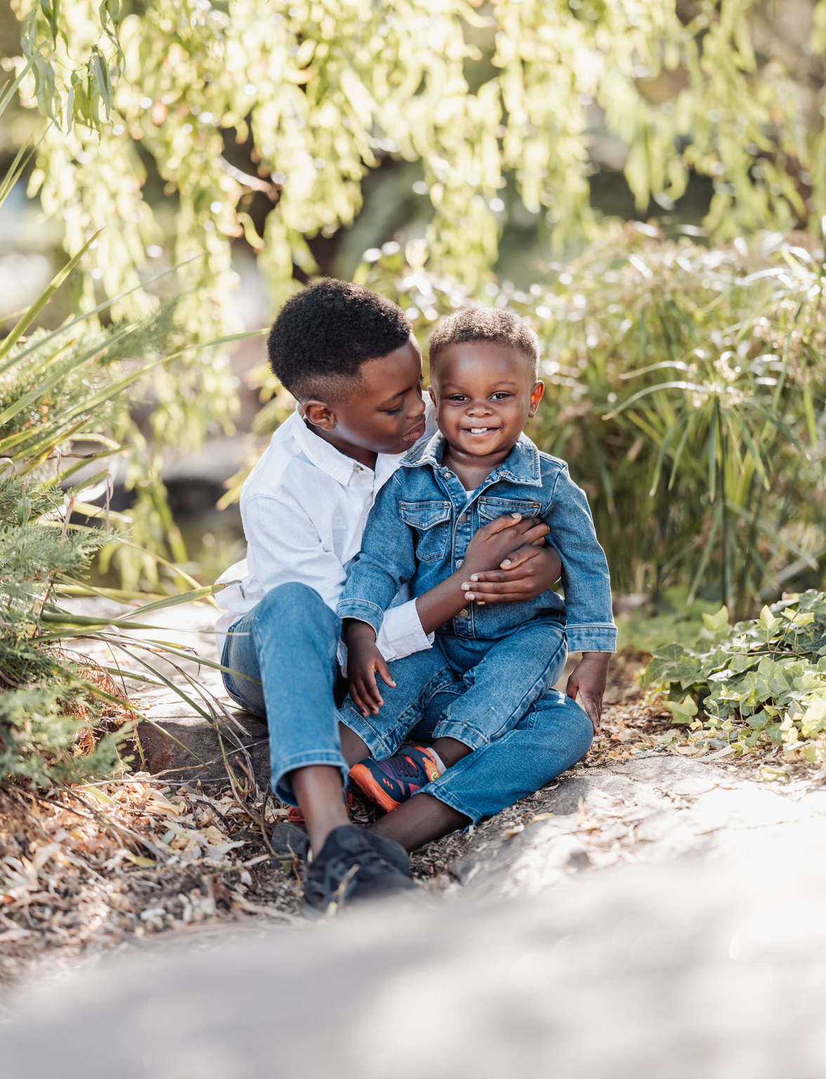 family photoshoot in veale gardens 1 Two young siblings bond joyfully outdoors, surrounded by greenery and natural sunlight.