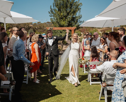 Newlyweds walk down the aisle holding hands as wedding guests celebrate beneath white umbrellas at Tiny House On The Prairie.