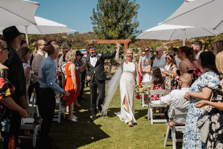 Newlyweds walk down the aisle holding hands as wedding guests celebrate beneath white umbrellas at Tiny House On The Prairie.