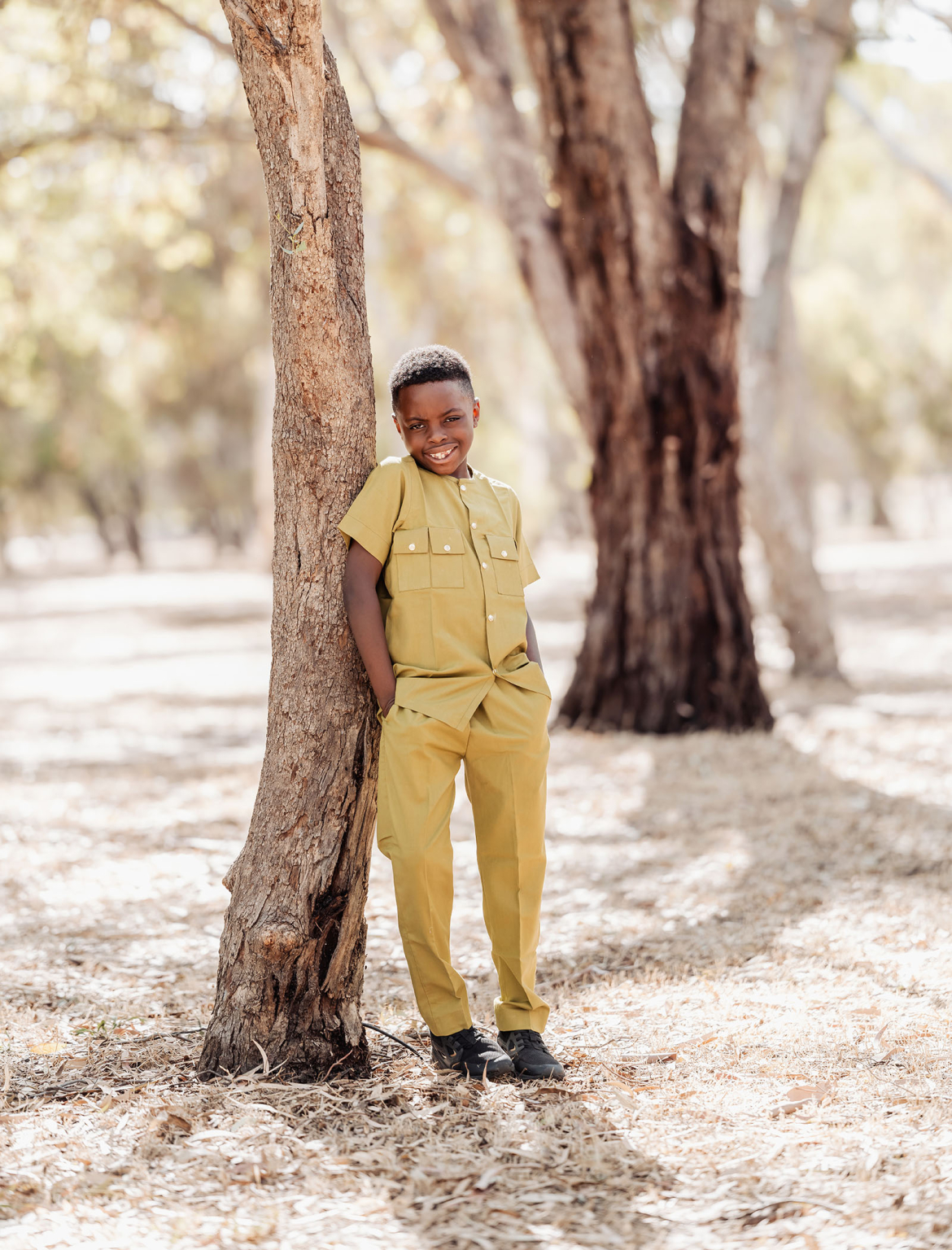family photoshoot in veale gardens 21 Young boy in light yellow outfit, leaning against a tree in a sunlit forest.