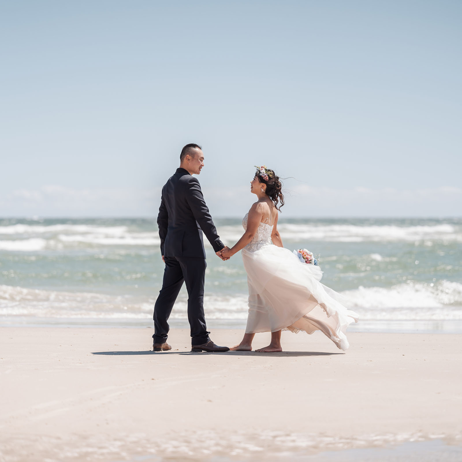 bumbunga lake photoshoot - tina & eric 38 Romantic couple holding hands on a beach, dressed in wedding attire with ocean backdrop.