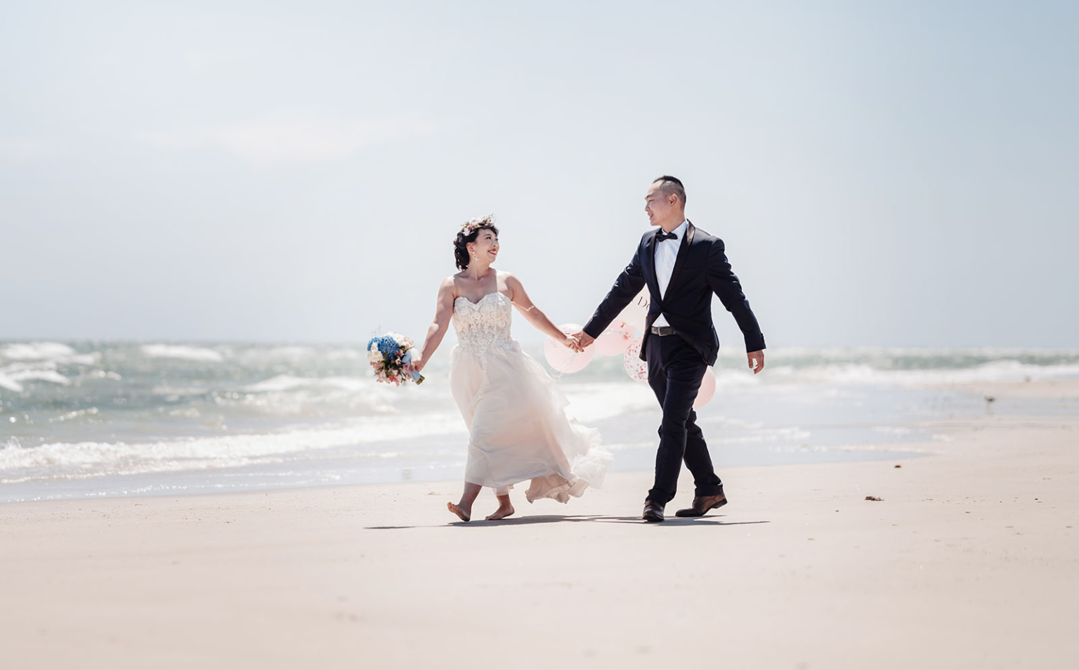 bumbunga lake photoshoot - tina & eric 32 Elegant couple in bridal attire walking on beach with flowers and balloons, celebrating love.