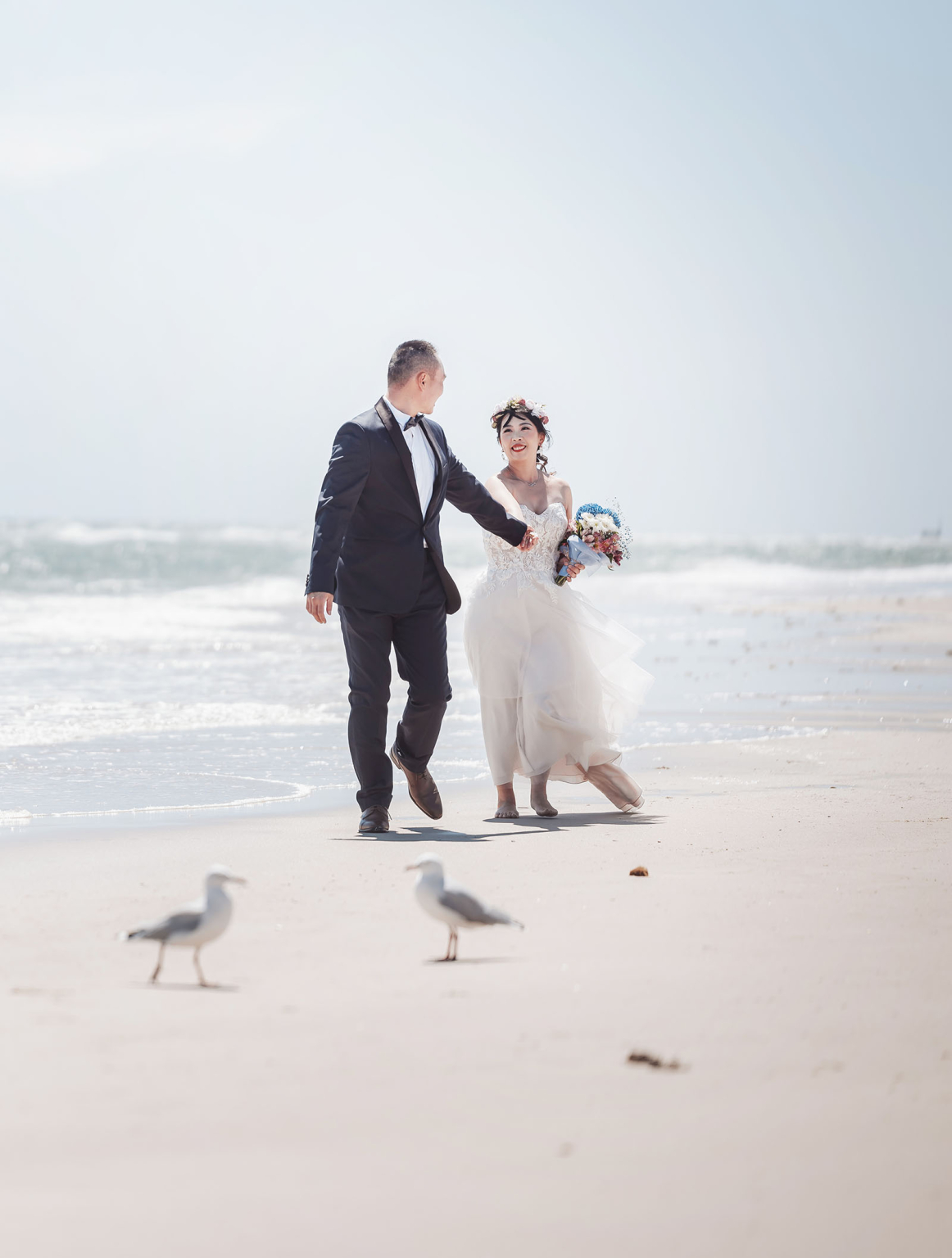 bumbunga lake photoshoot - tina & eric 36 Wedding couple walks on serene beach; bride in gown, groom in suit, ocean backdrop.