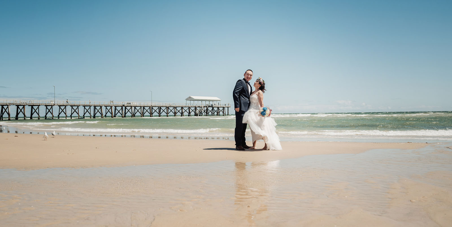bumbunga lake photoshoot - tina & eric 37 Beach wedding couple with ocean backdrop, wooden pier, and seagulls under blue sky.