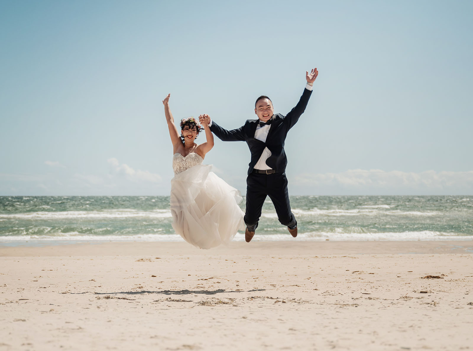 bumbunga lake photoshoot - tina & eric 46 Joyful couple jumping on beach in wedding attire under clear blue sky.