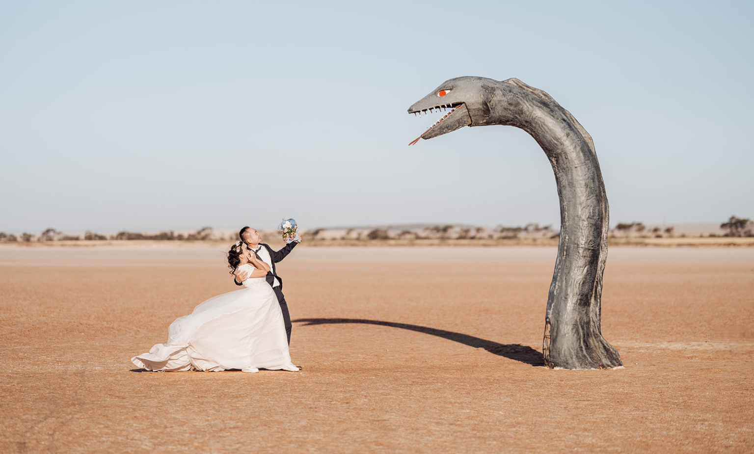 bumbunga lake photoshoot - tina & eric 77 Desert wedding couple poses near large serpent sculpture under blue sky.
