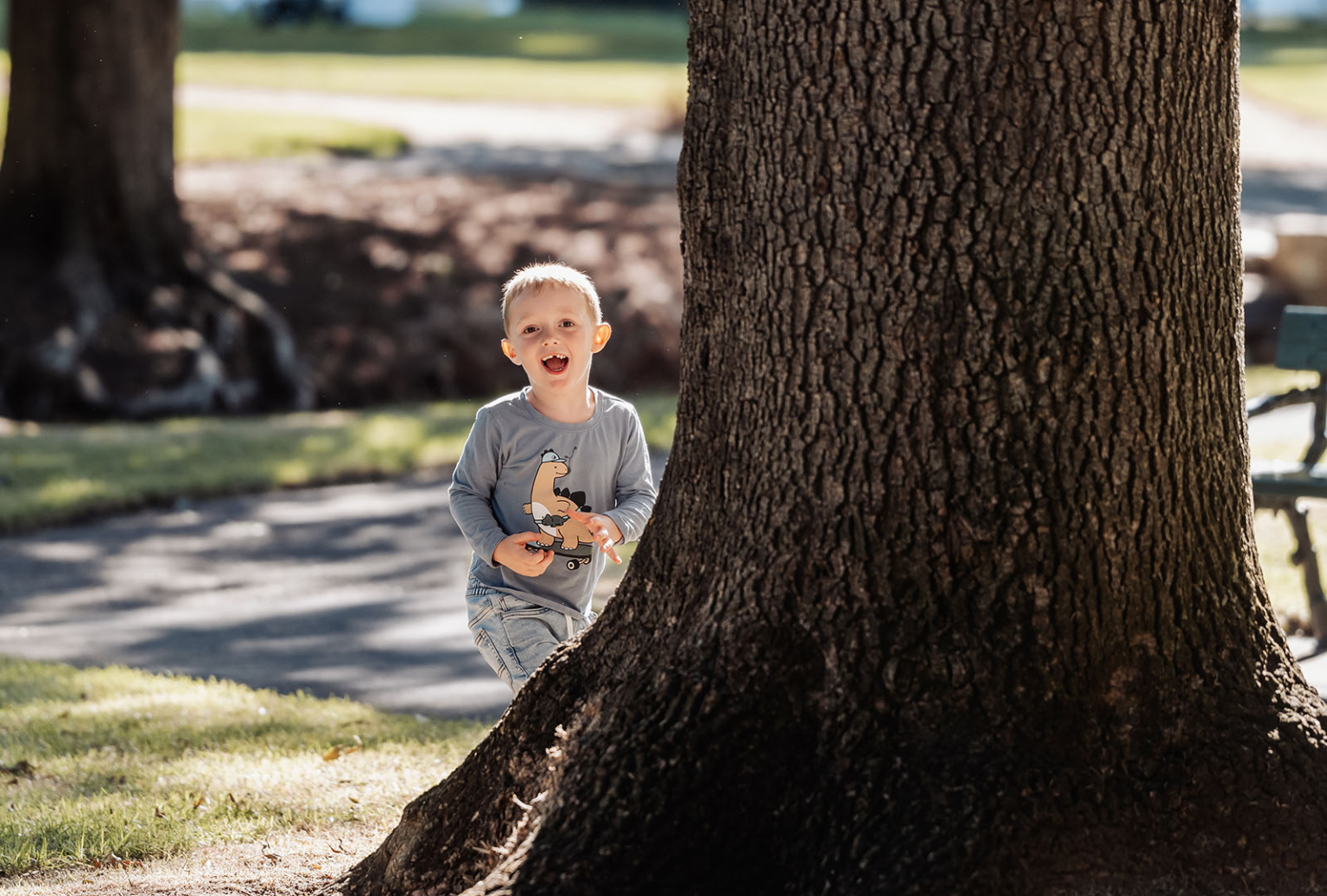 veale gardens family photography 106 Child running joyfully by tree in sunlit park with vibrant grass and pathway.