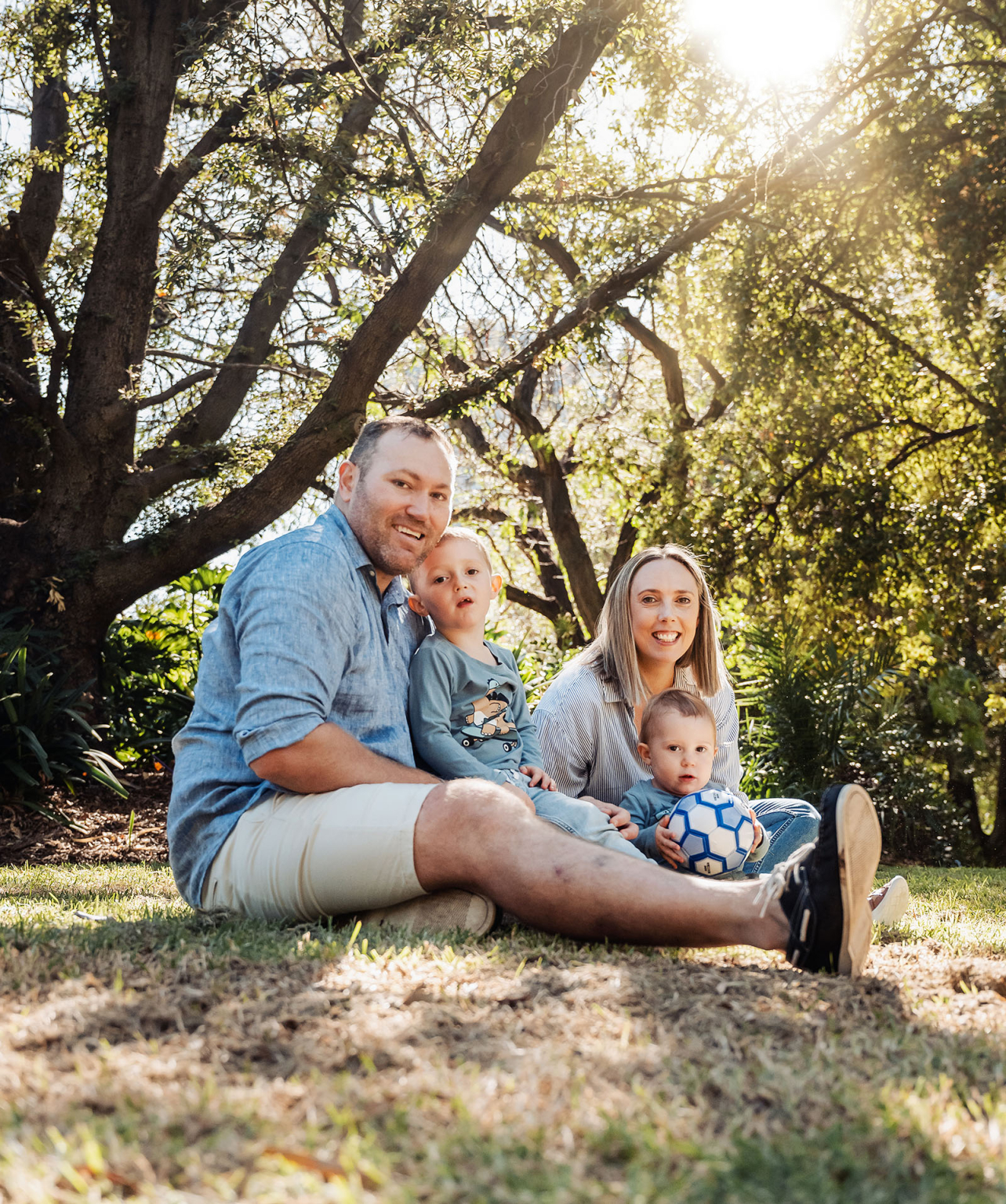veale gardens family photography 119 Family enjoying sunny day outdoors, sitting on grass with soccer ball under tree.