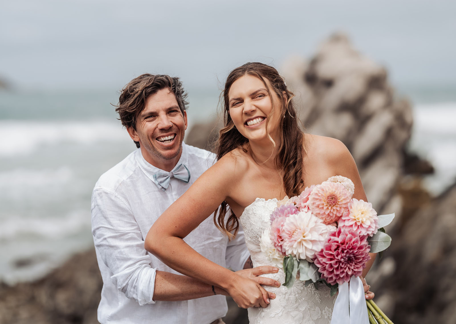 the many benefits of hiring professional wedding photographers 2 Joyful couple laughing on rocky beach, wedding attire, ocean waves in background.