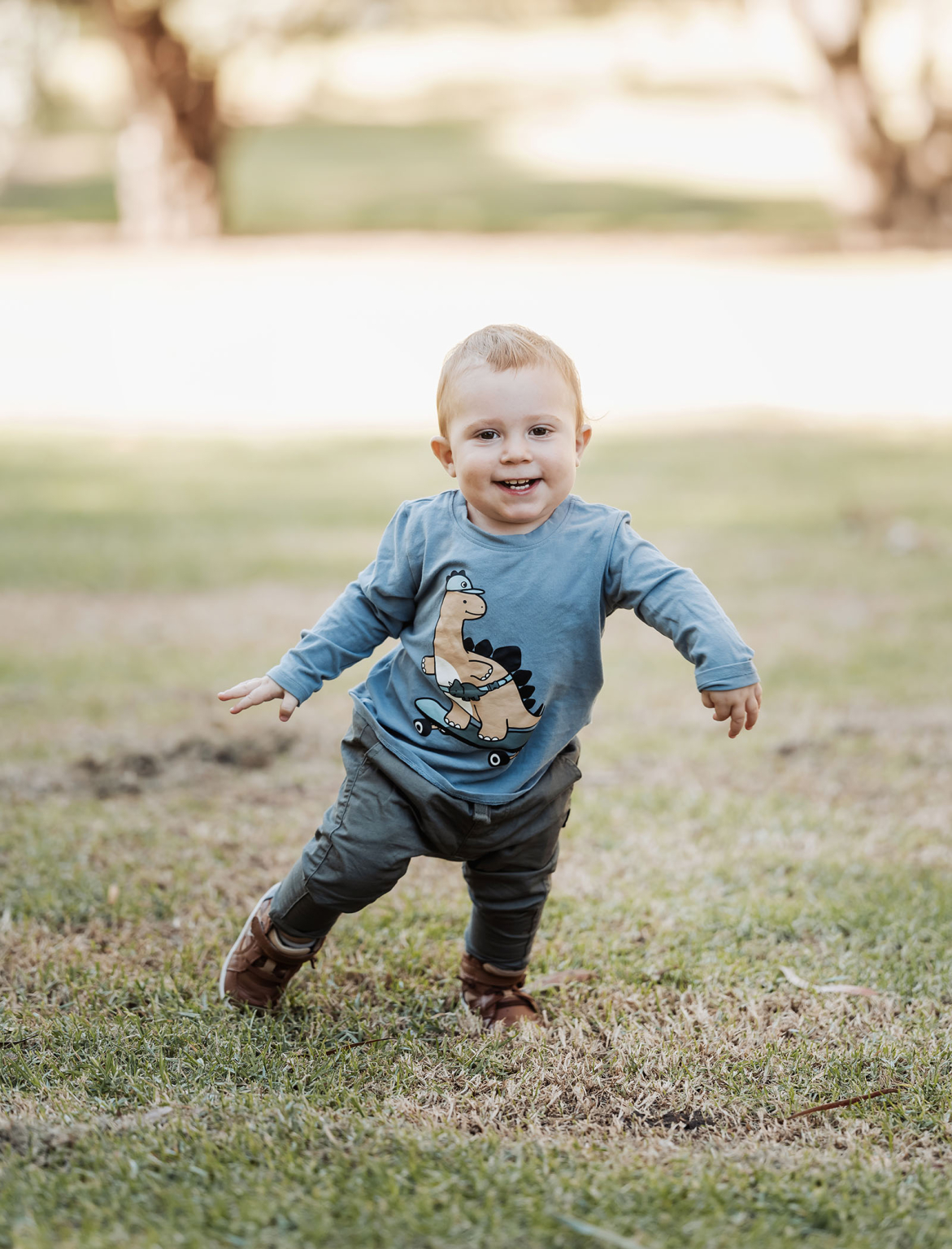 veale gardens family photography 115 A joyful child in a blue dinosaur shirt walks in a sunny grassy park.
