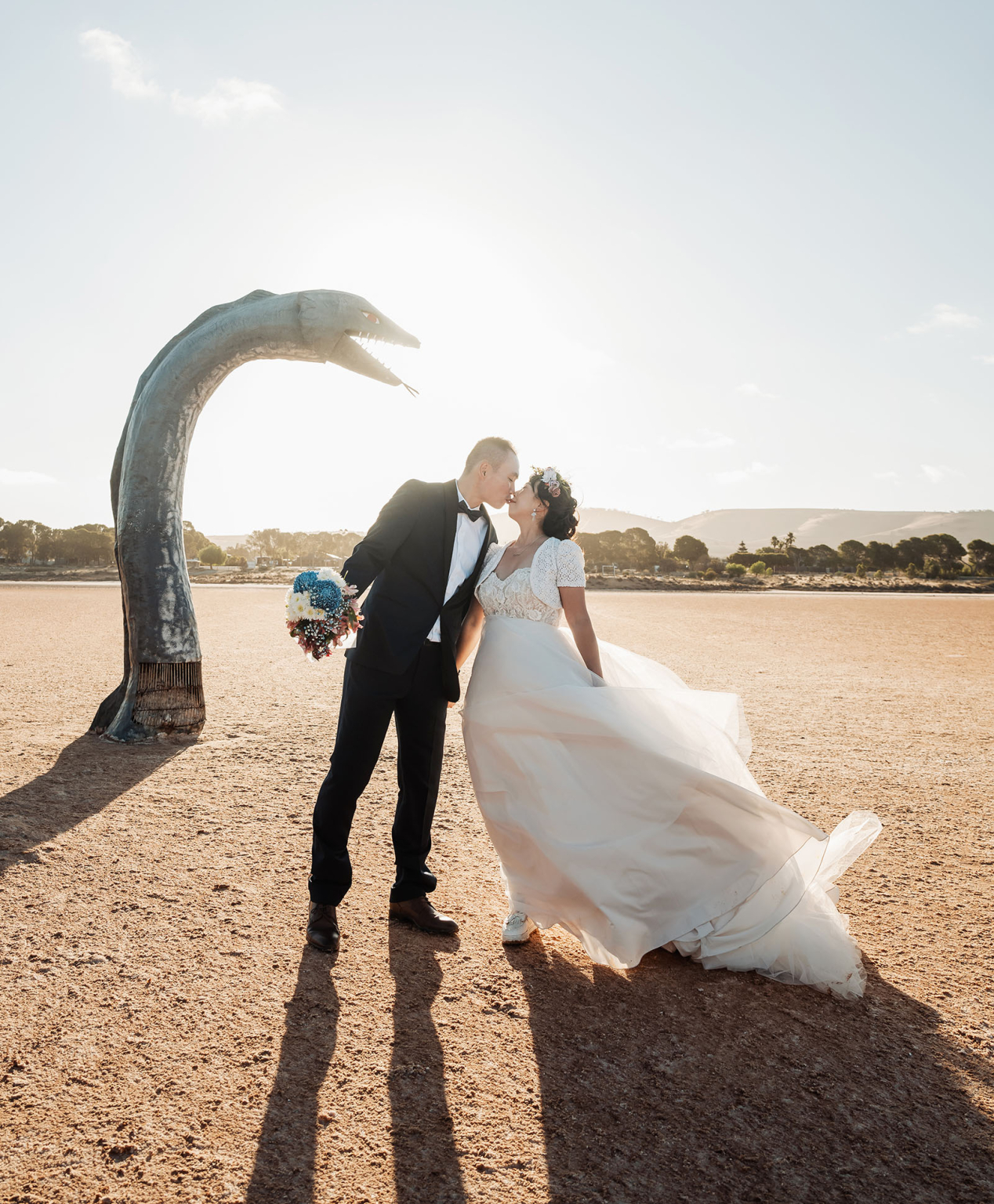 bumbunga lake photoshoot - tina & eric 79 A couple kisses at sunset, framed by a whimsical sculpture in a picturesque outdoor setting.