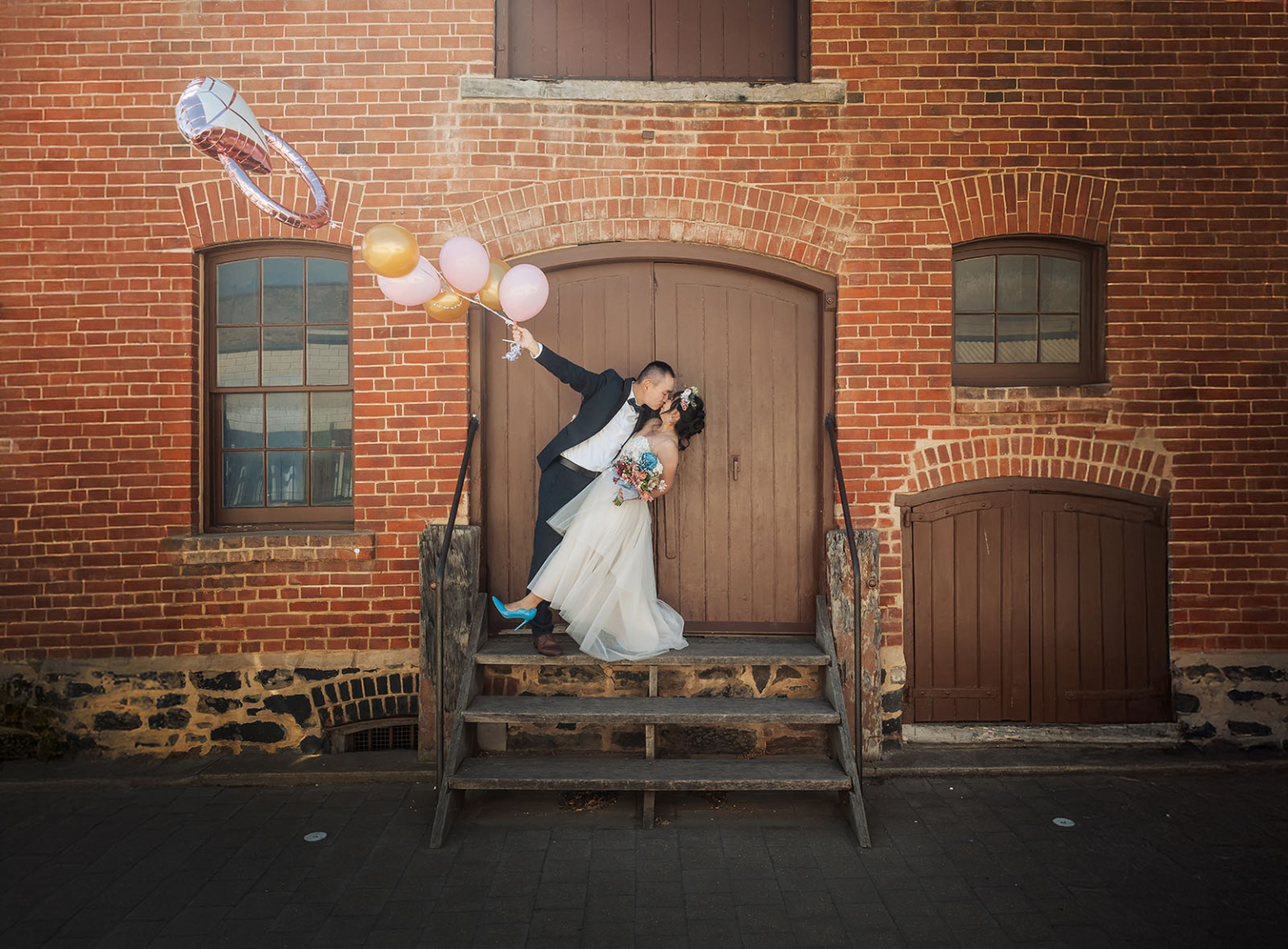 bumbunga lake photoshoot - tina & eric 55 Newlywed couple kissing on stairs with balloons and rustic brick building background.