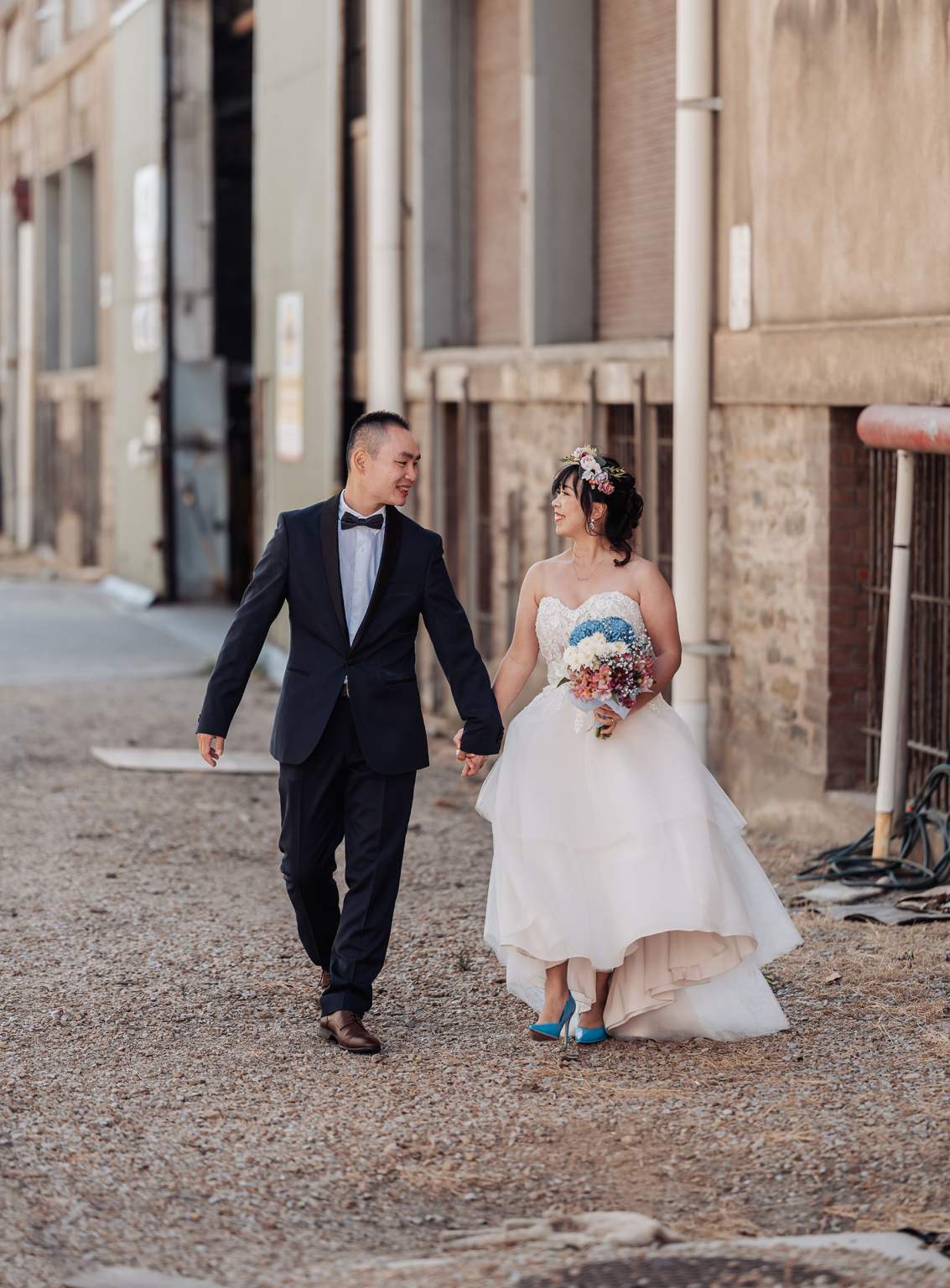 bumbunga lake photoshoot - tina & eric 49 Wedding couple walking joyfully on rustic path, wearing stylish attire, surrounded by industrial buildings.