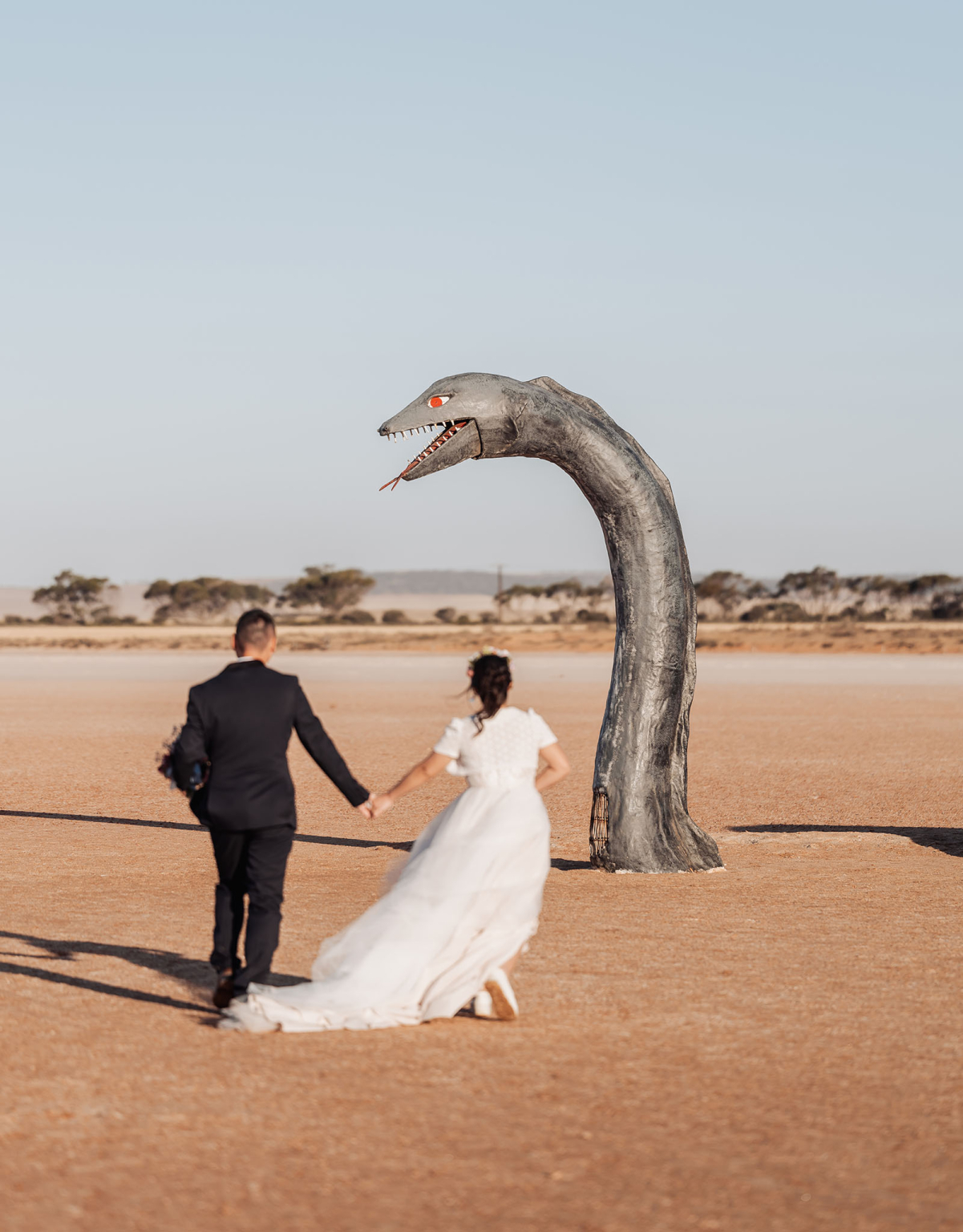 bumbunga lake photoshoot - tina & eric 76 Bride and groom in desert with towering serpent-like sculpture under clear blue sky.
