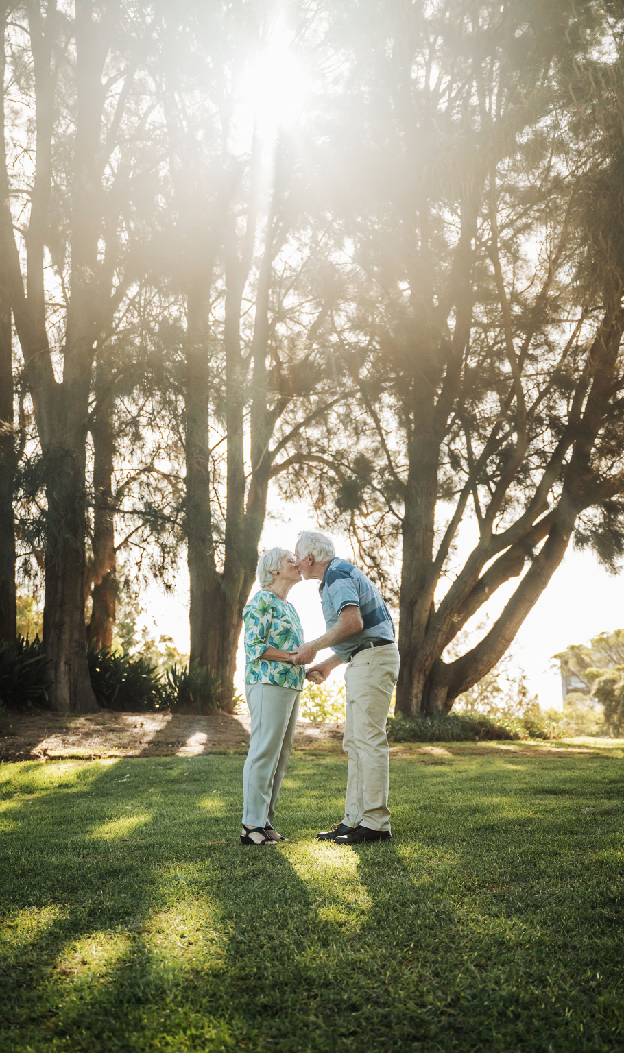 veale gardens family photoshoot 19 Elderly couple sharing a kiss in sunlit park surrounded by lush greenery and tall trees.