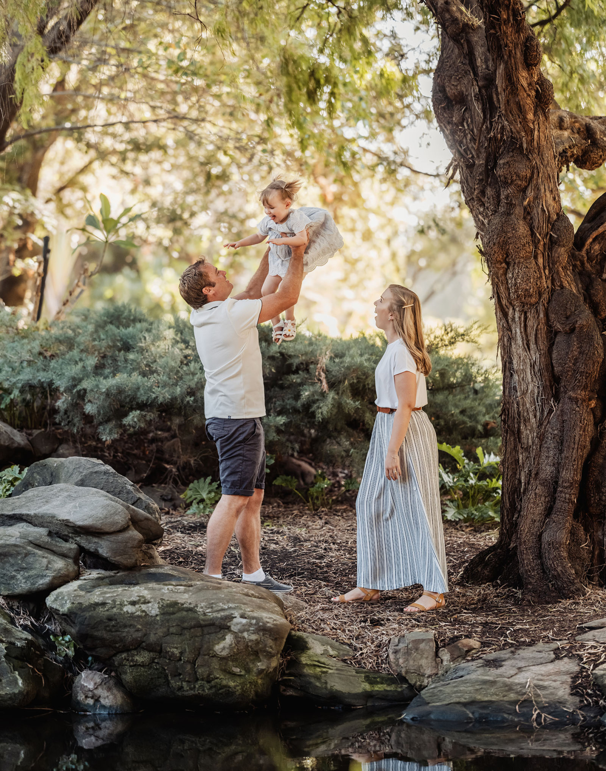 veale gardens family photoshoot 8 Family enjoying nature: father lifting daughter, mother smiling, amidst trees and sunlight.
