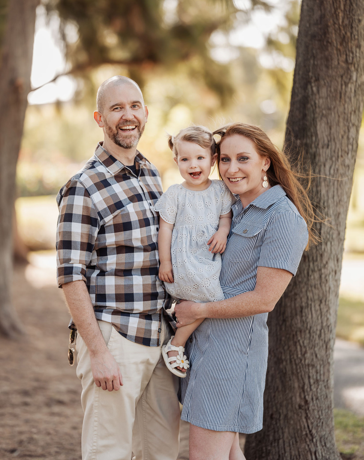 veale gardens family photoshoot 12 Family enjoying a sunny day outdoors, parents holding their smiling child near a tree.