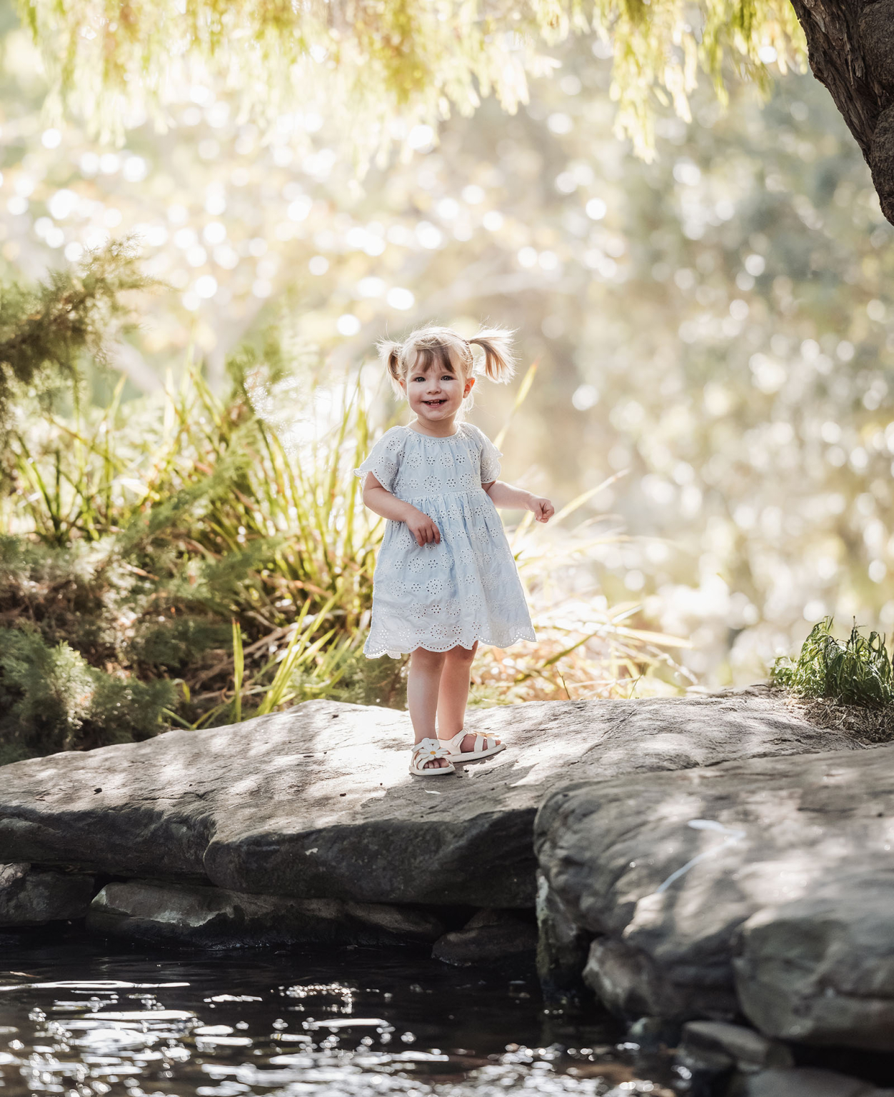 veale gardens family photoshoot 2 Child in white dress on rock by stream, surrounded by lush greenery and sunlight.