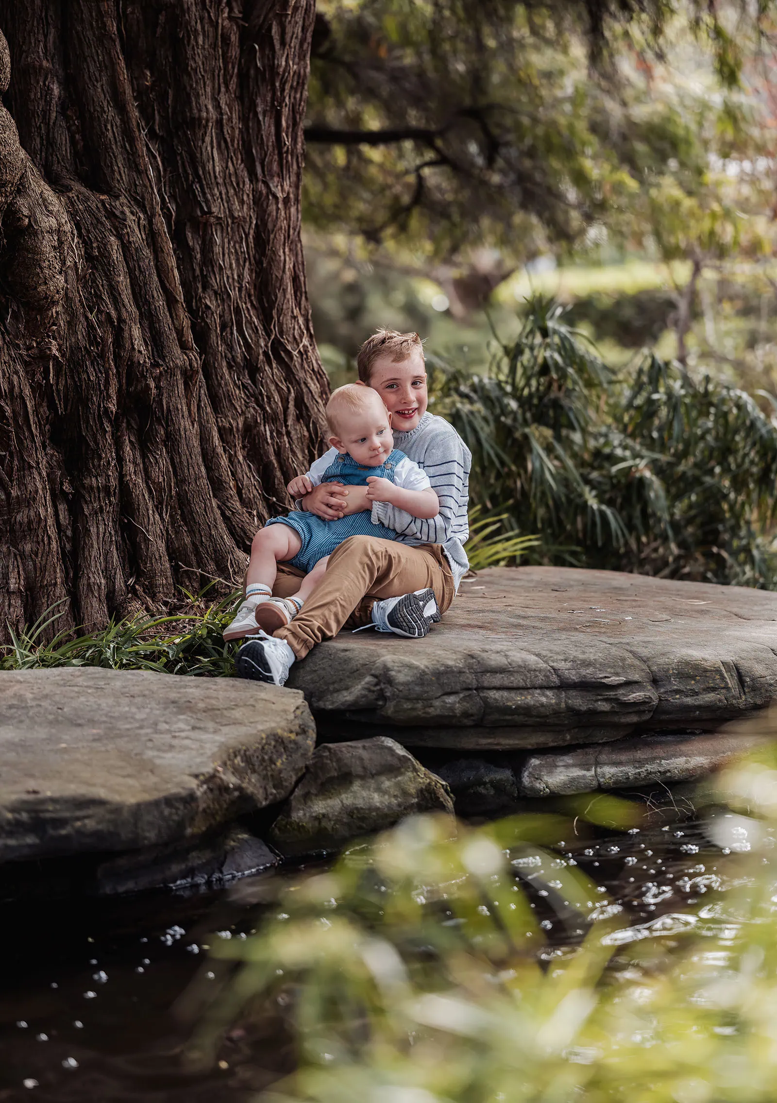 southern parklands family photography 21 Two siblings smiling together under a large tree in a sunny park.