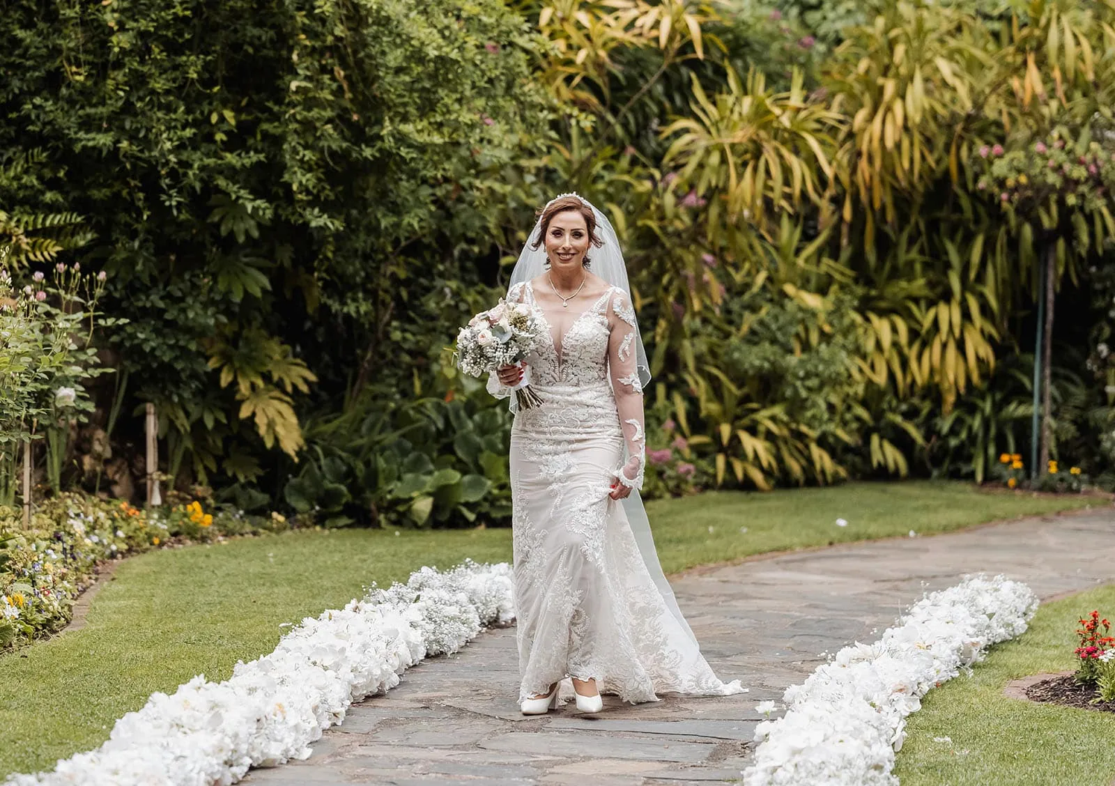 wedding at the manor in basket range - maryam & jason 132 Bride walking along a picturesque garden pathway, surrounded by flowers and lush greenery.