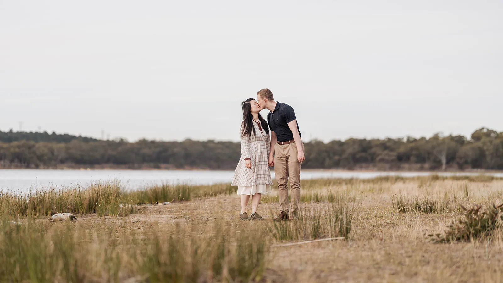 happy valley reservoir family shoot 103 Couple kissing by a serene lake, under a cloudy sky, surrounded by nature.