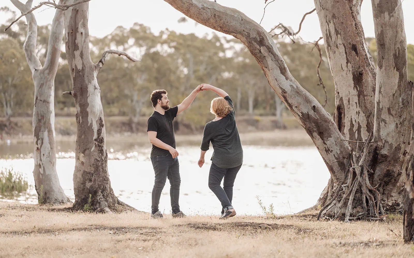 happy valley reservoir family shoot 95 Couple joyfully dancing by water, surrounded by eucalyptus trees, during a serene, warm evening.