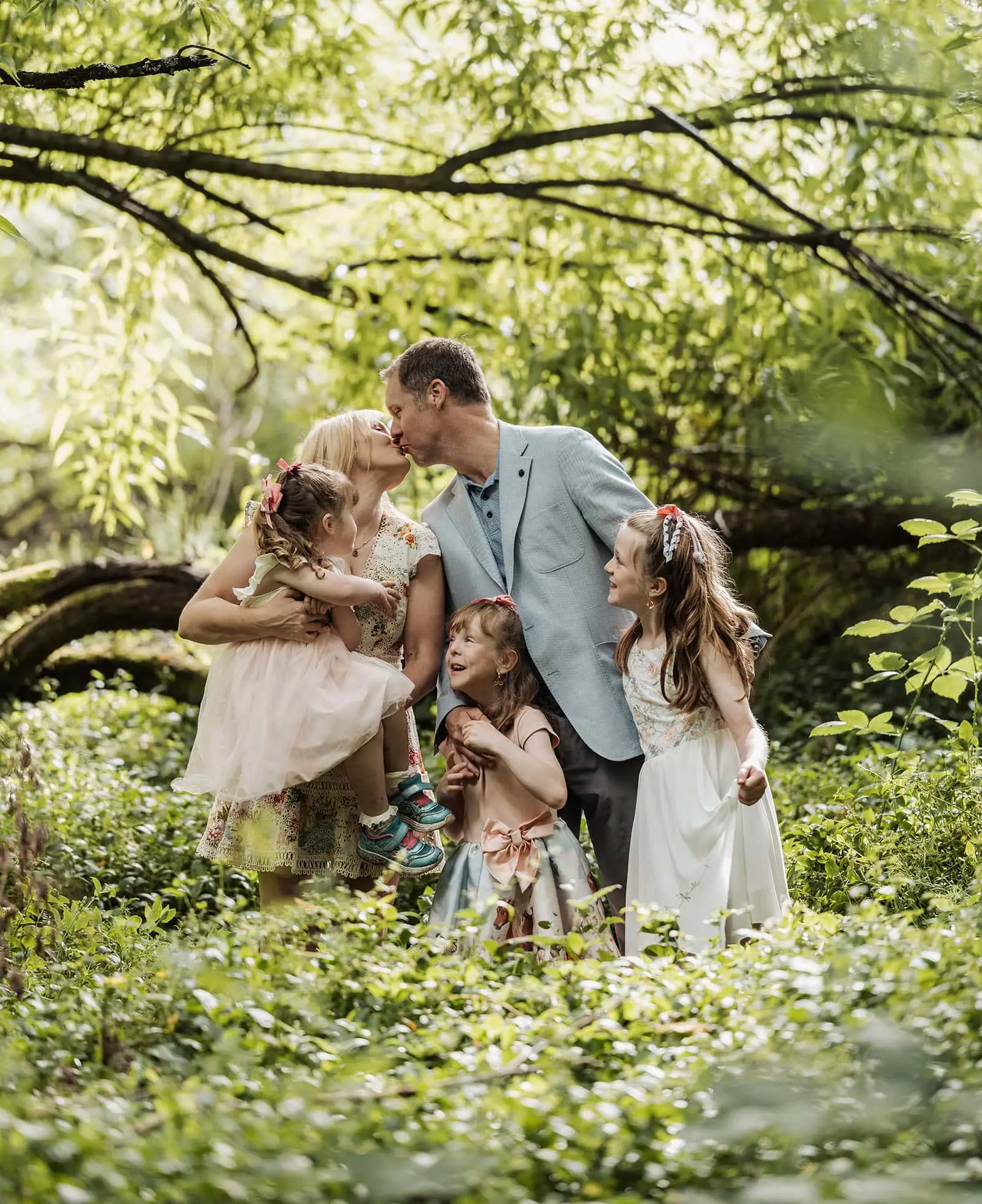 ironbank family photography 117 Loving family in nature, parents kissing, daughters in vibrant dresses, joyful and harmonious moment.