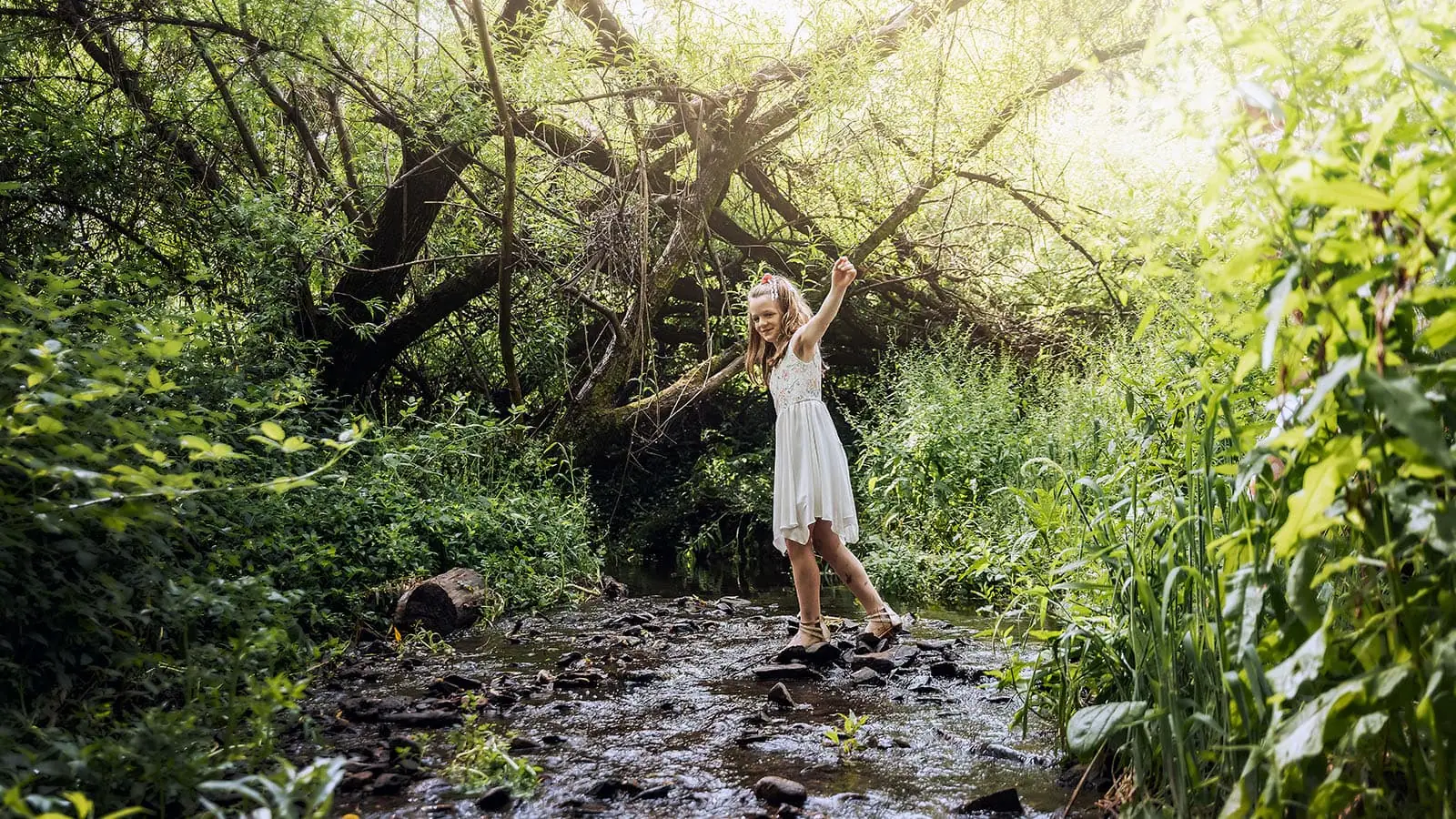 ironbank family photography 130 Young girl joyfully balances on rocks in a sunlit forest stream, surrounded by lush greenery.