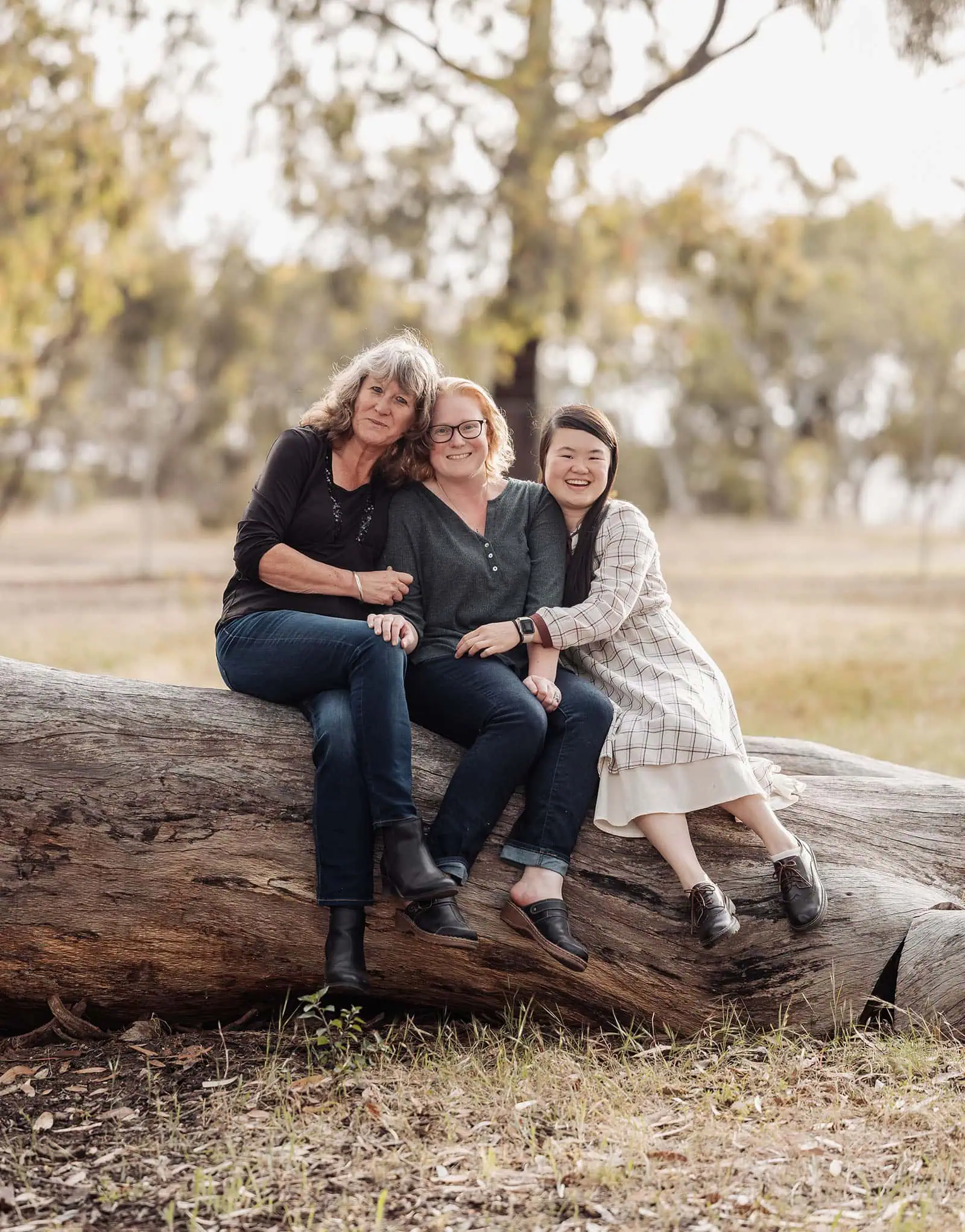 happy valley reservoir family shoot 85 Three women smiling on a fallen log in a lush outdoor setting.