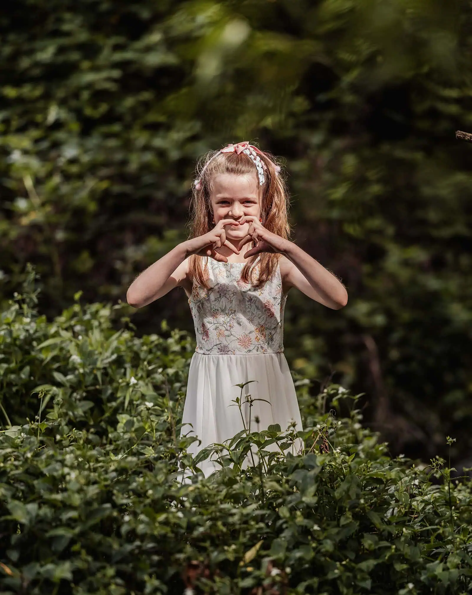 ironbank family photography 124 Young girl in white dress surrounded by lush greenery forming heart with hands.