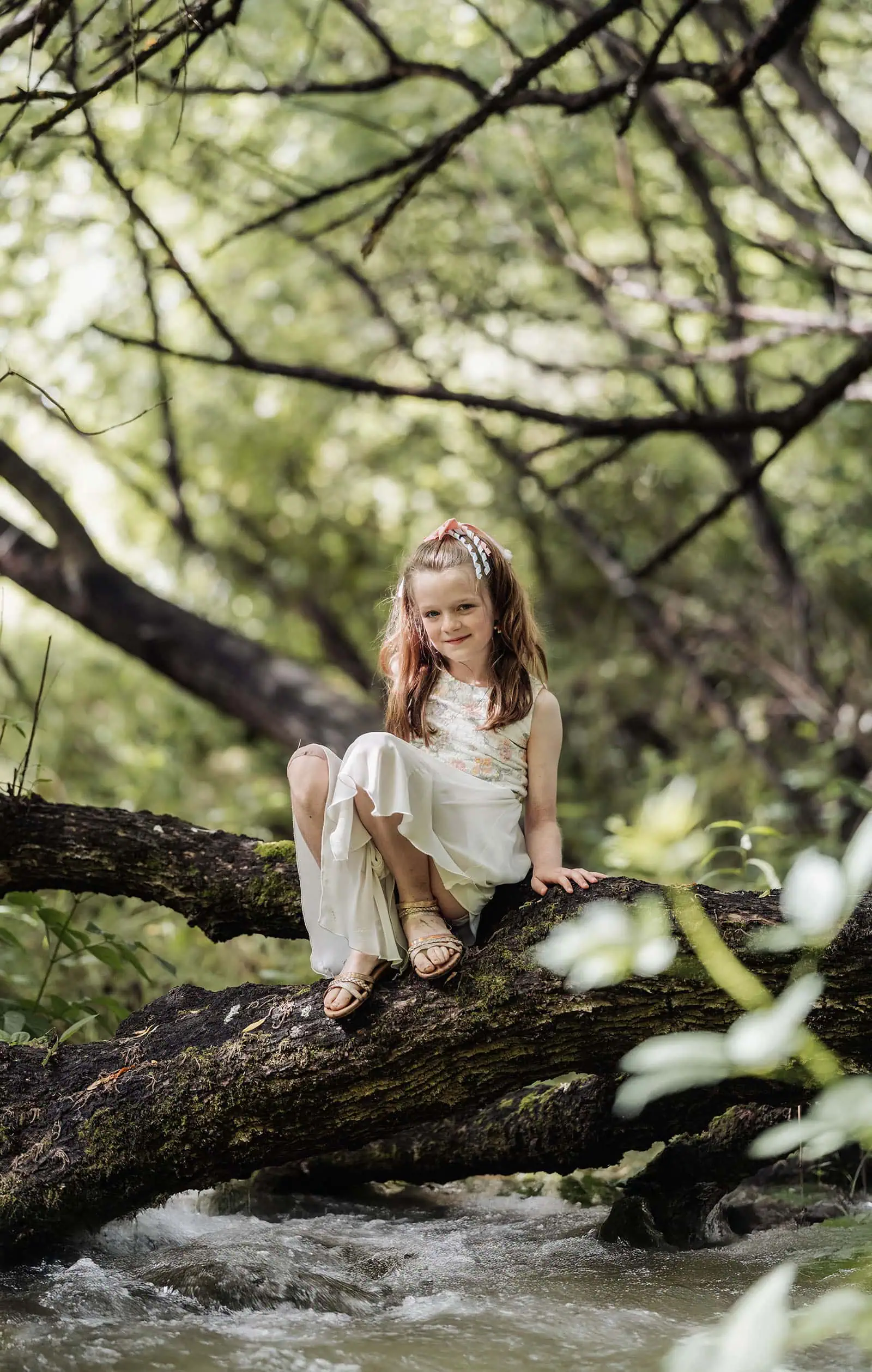 ironbank family photography 128 Young girl in floral dress sitting on mossy tree branch over stream in lush forest.