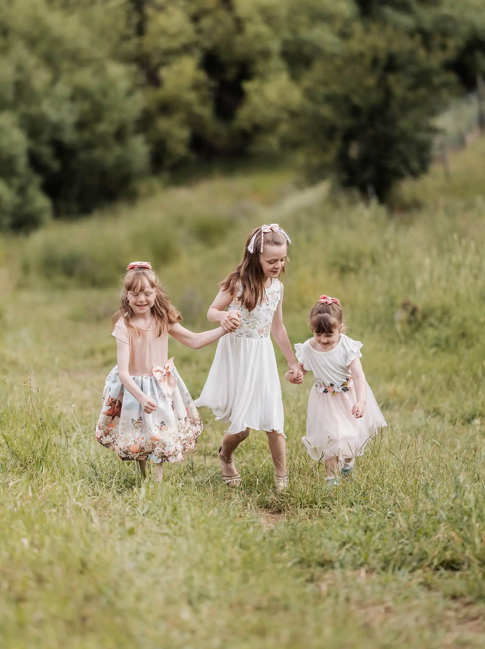ironbank family photography 110 Three young girls in floral summer dresses joyfully walk hand-in-hand through a lush green meadow.