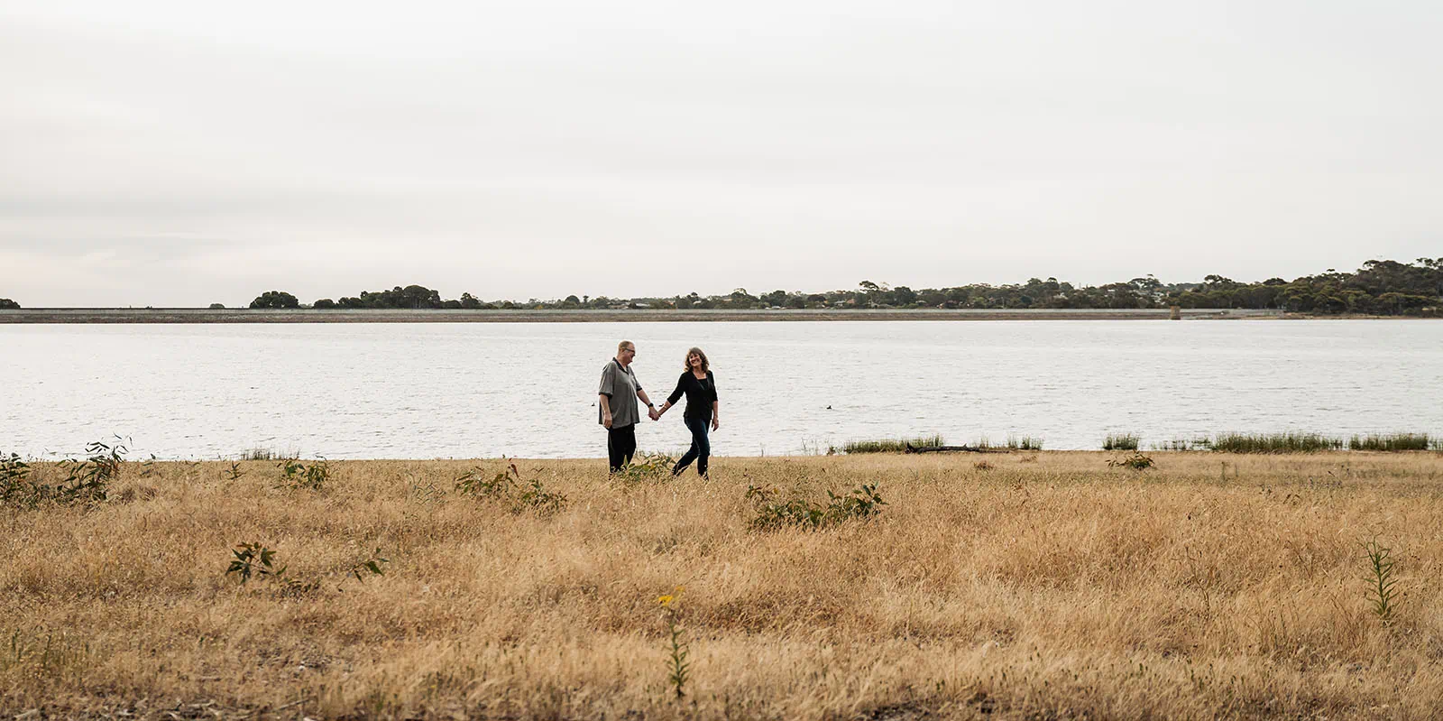 happy valley reservoir family shoot 100 Couple walking on grassy field by tranquil water under overcast sky.