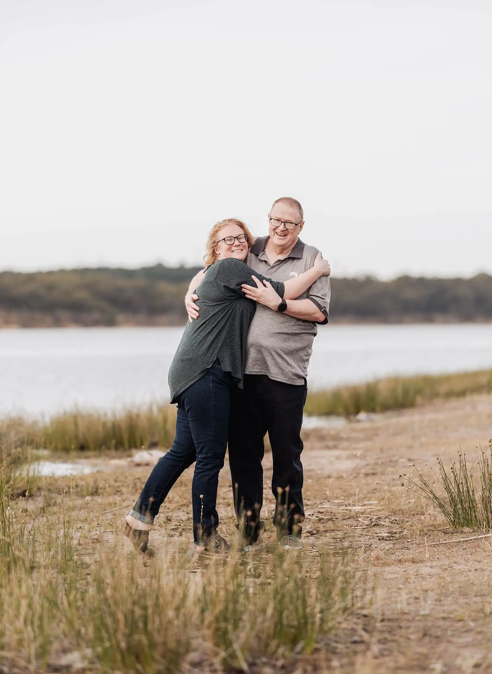 happy valley reservoir family shoot 105 Two people embrace joyfully by a serene lake under an overcast sky.