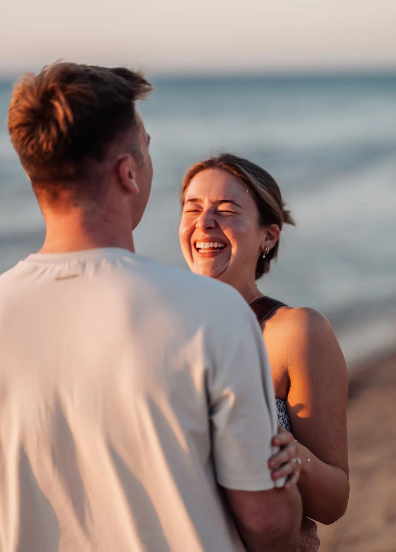 christies beach proposal – hollie & william 43 Joyful couple laughing on beach during sunset, ocean waves in background.