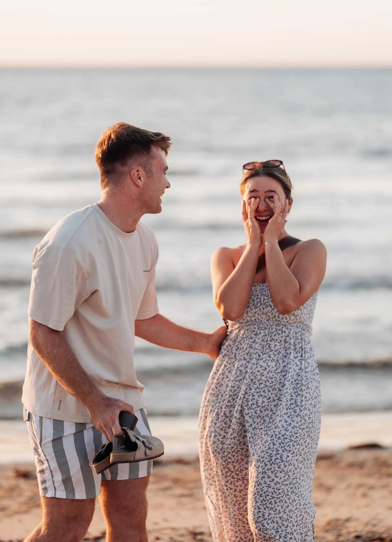 christies beach proposal – hollie & william 38 Joyful couple on sandy beach with ocean waves and warm sunset glow.