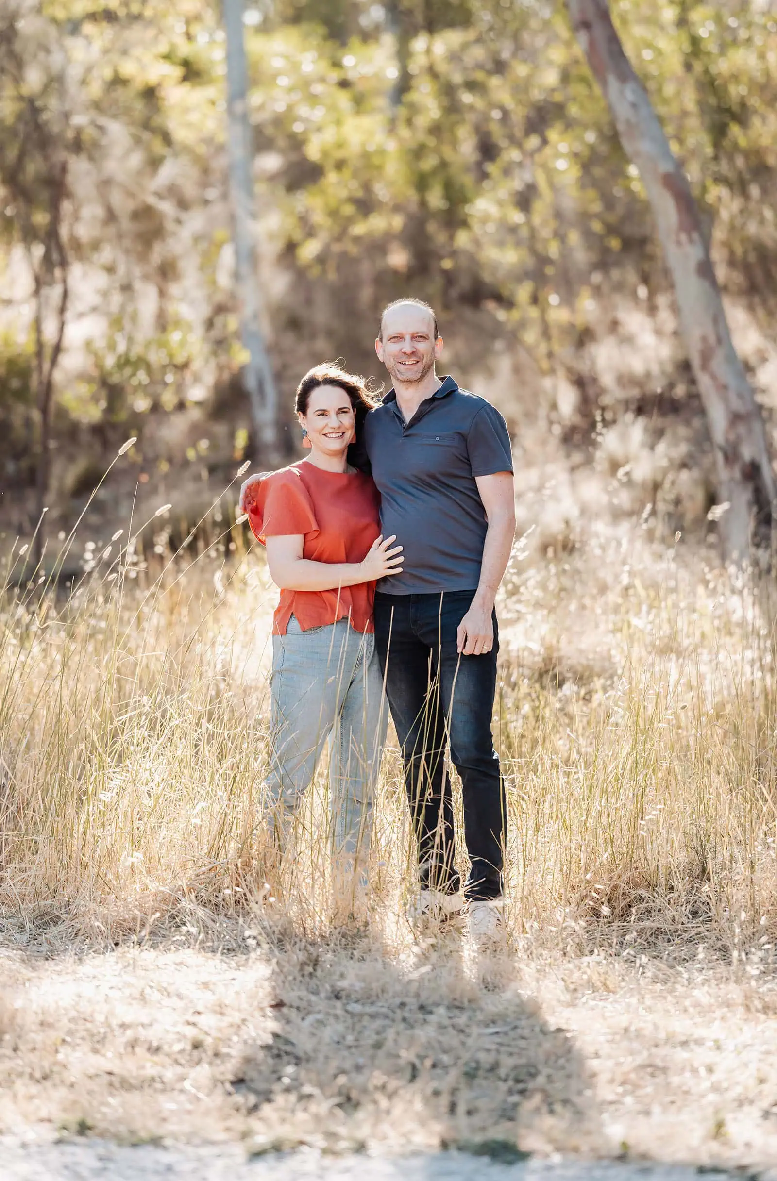 beautiful anstey hill family photoshoot 3 Couple embracing in a sunlit field with trees, wearing casual orange and blue attire.