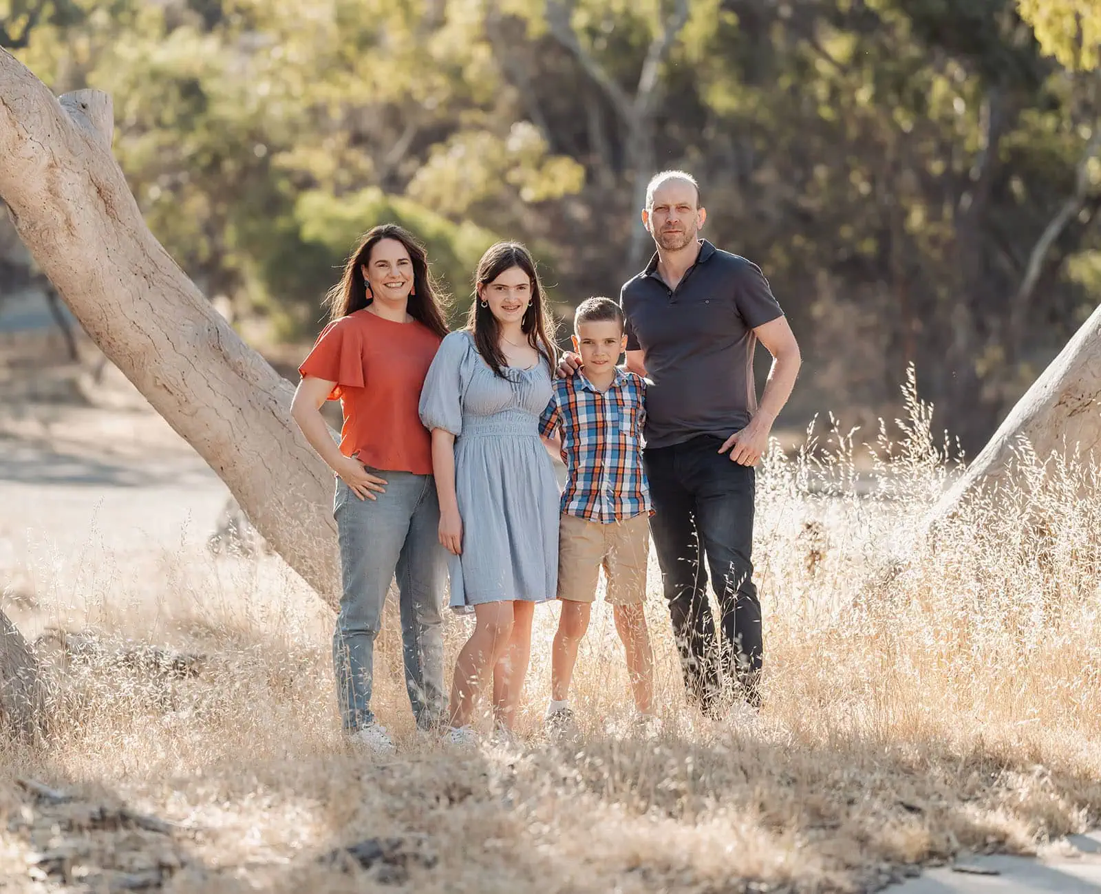 beautiful anstey hill family photoshoot 11 Family posing warmly in a serene, sunlit outdoor setting with trees and dry grass.