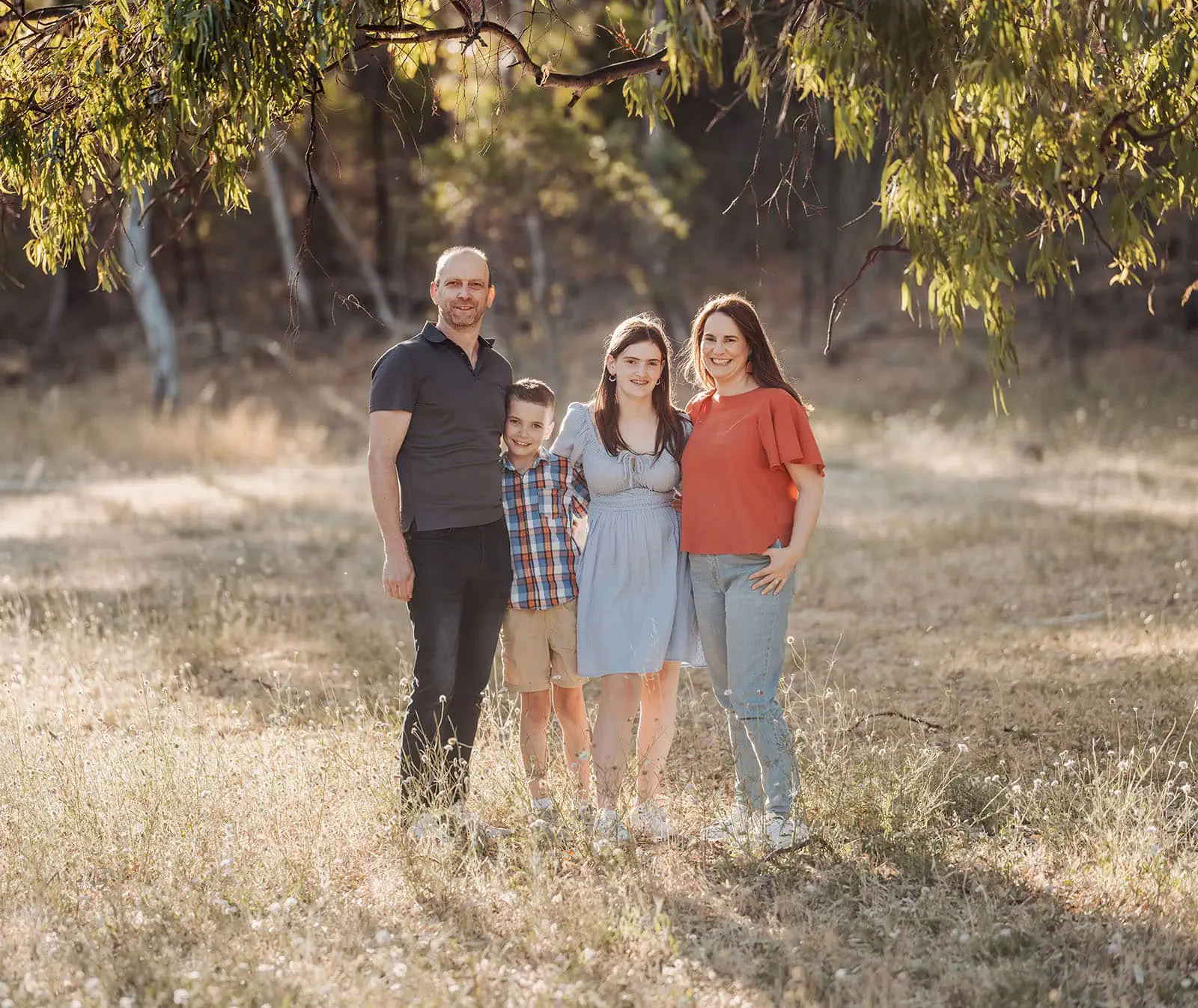 beautiful anstey hill family photoshoot 26 Family of four smiling in a sunlit field with trees and flowers.
