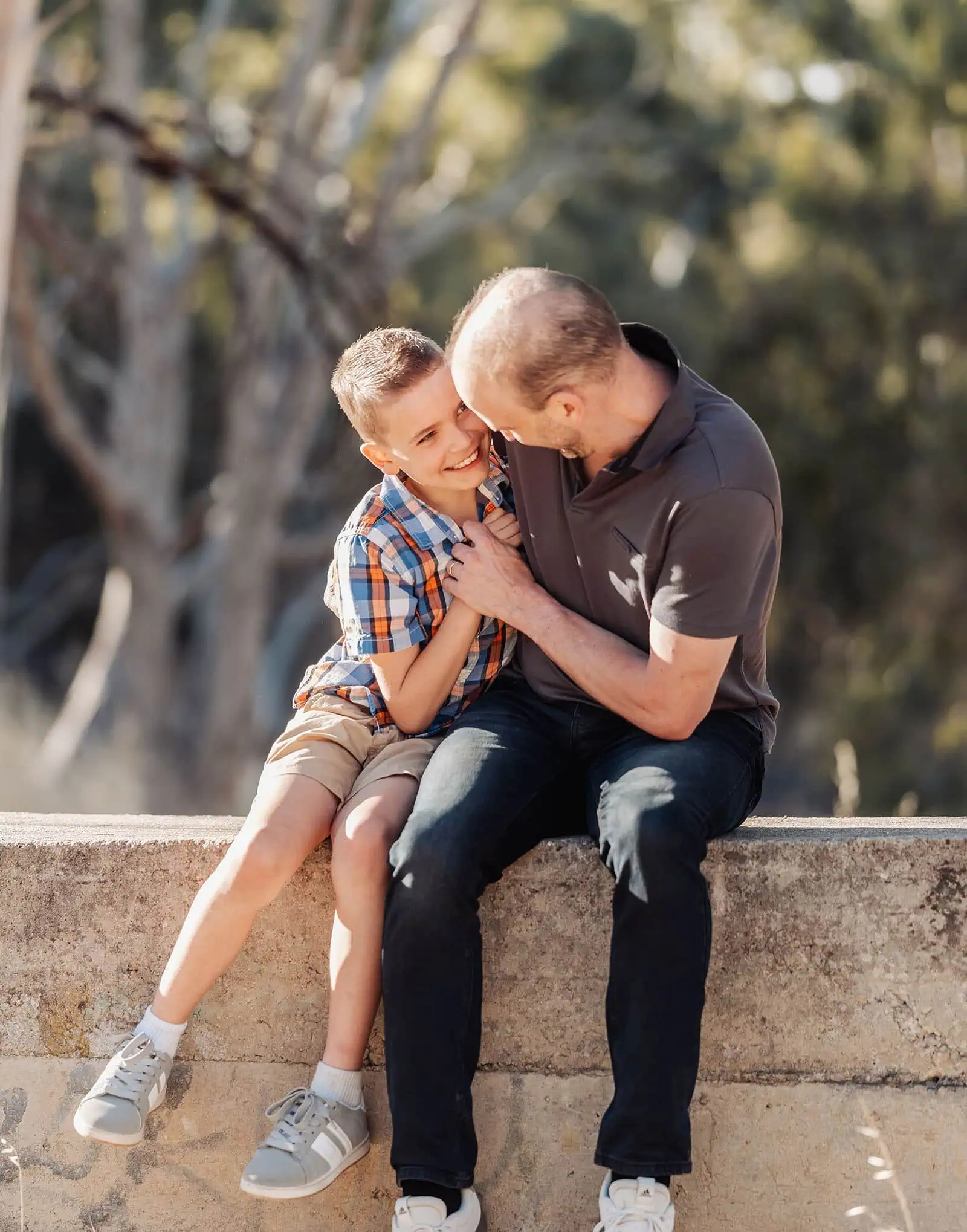 beautiful anstey hill family photoshoot 16 Father and son share a joyful moment sitting outdoors on stone ledge.