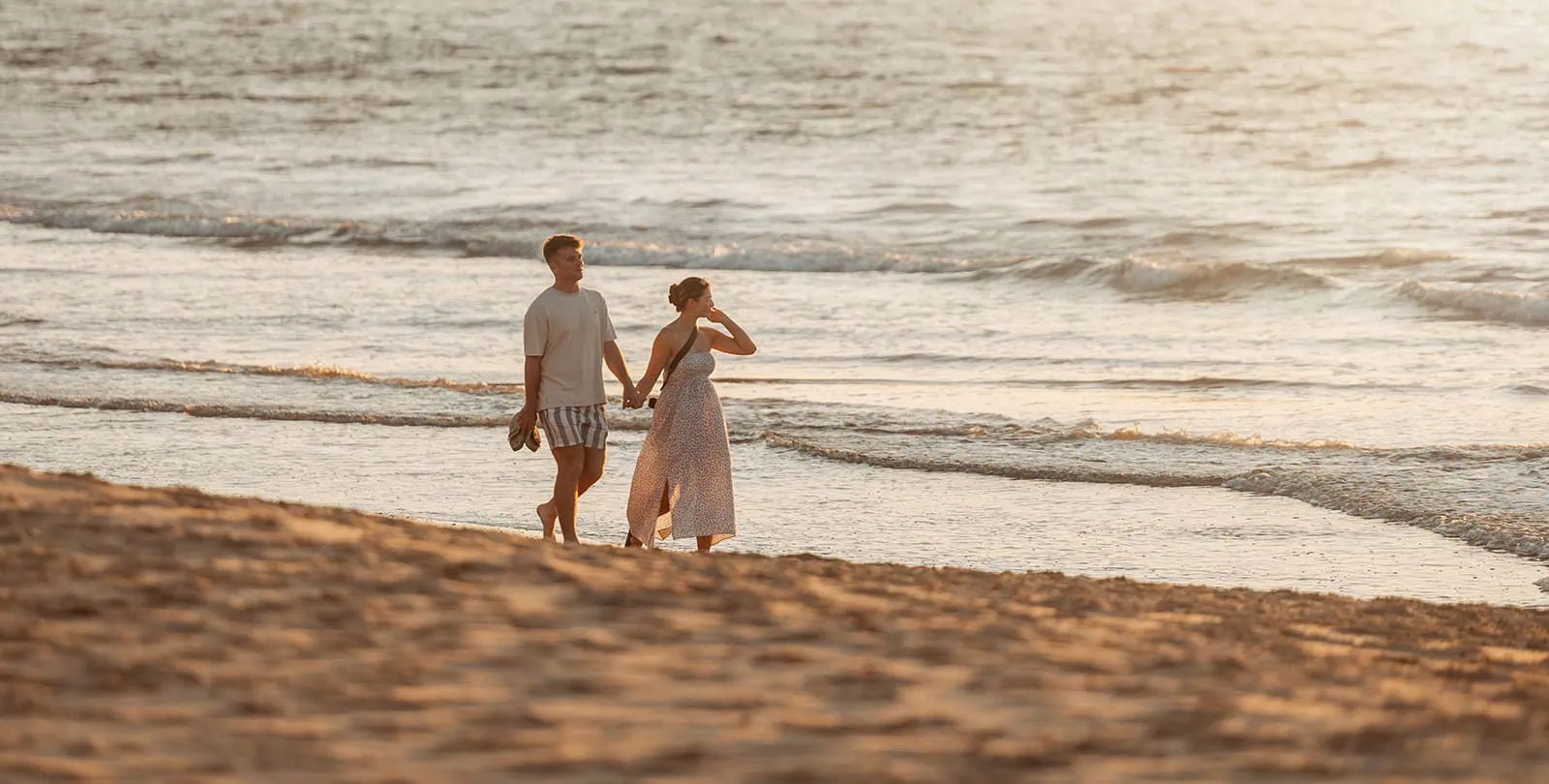christies beach proposal – hollie & william 30 Couple strolling on serene beach during golden hour, with warm sunset hues reflecting on water.