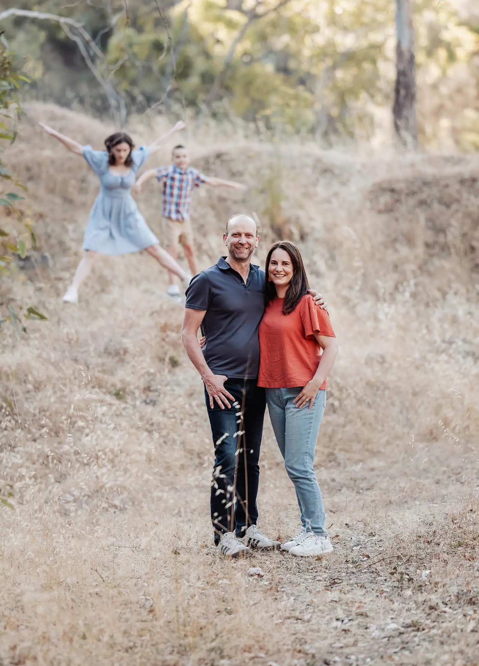beautiful anstey hill family photoshoot 14 Joyful couple standing in sunlit field with children playing in background.