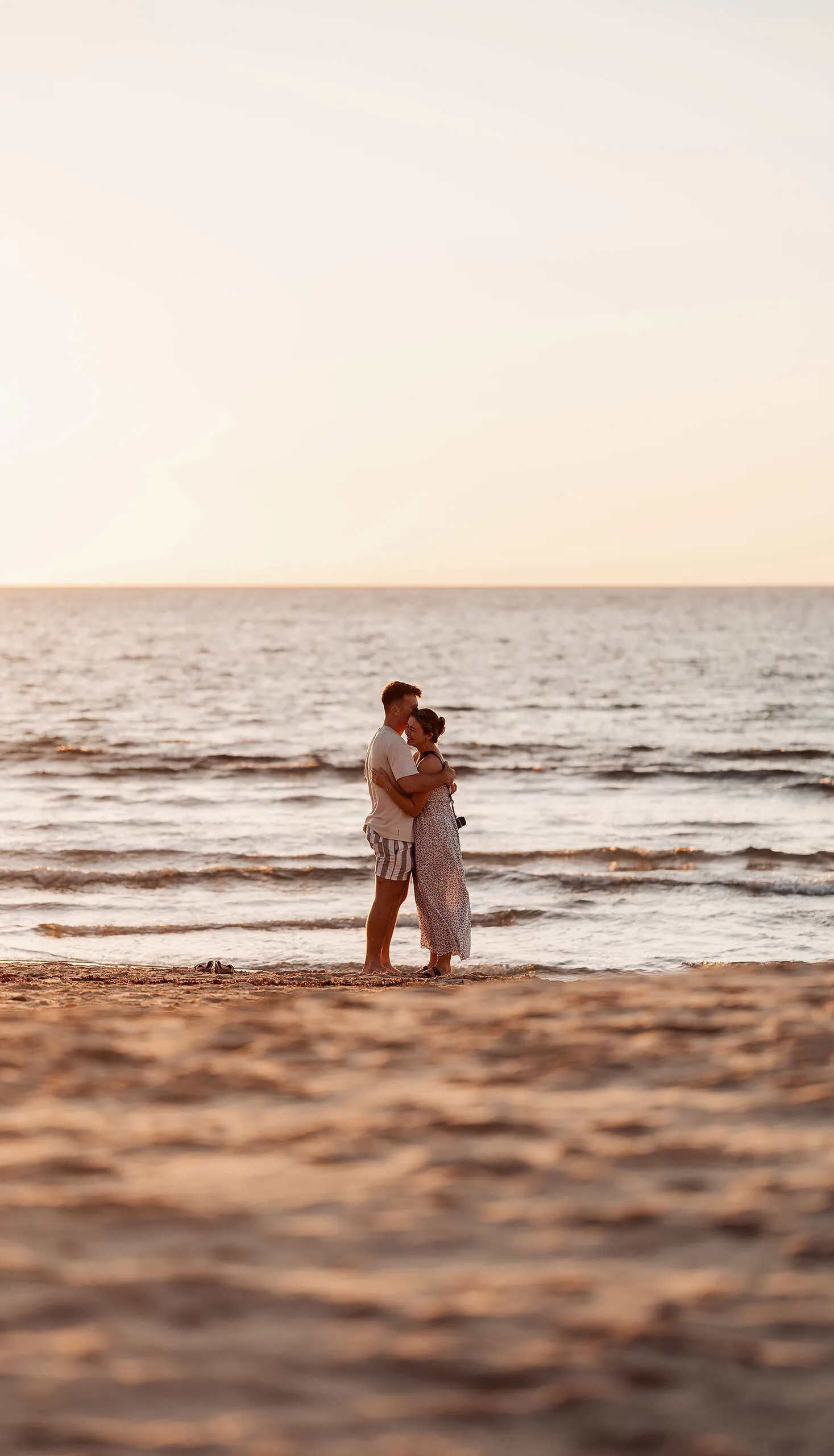 christies beach proposal – hollie & william 36 Couple embracing on a serene beach at sunset, golden hour, calm ocean in background.