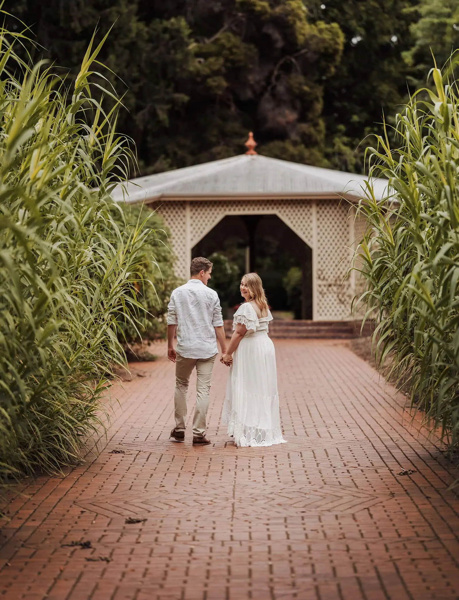 botanic gardens wedding shoot - olivia & sean 18 Couple strolling on brick path amid lush garden towards gazebo, exuding romance and tranquility.