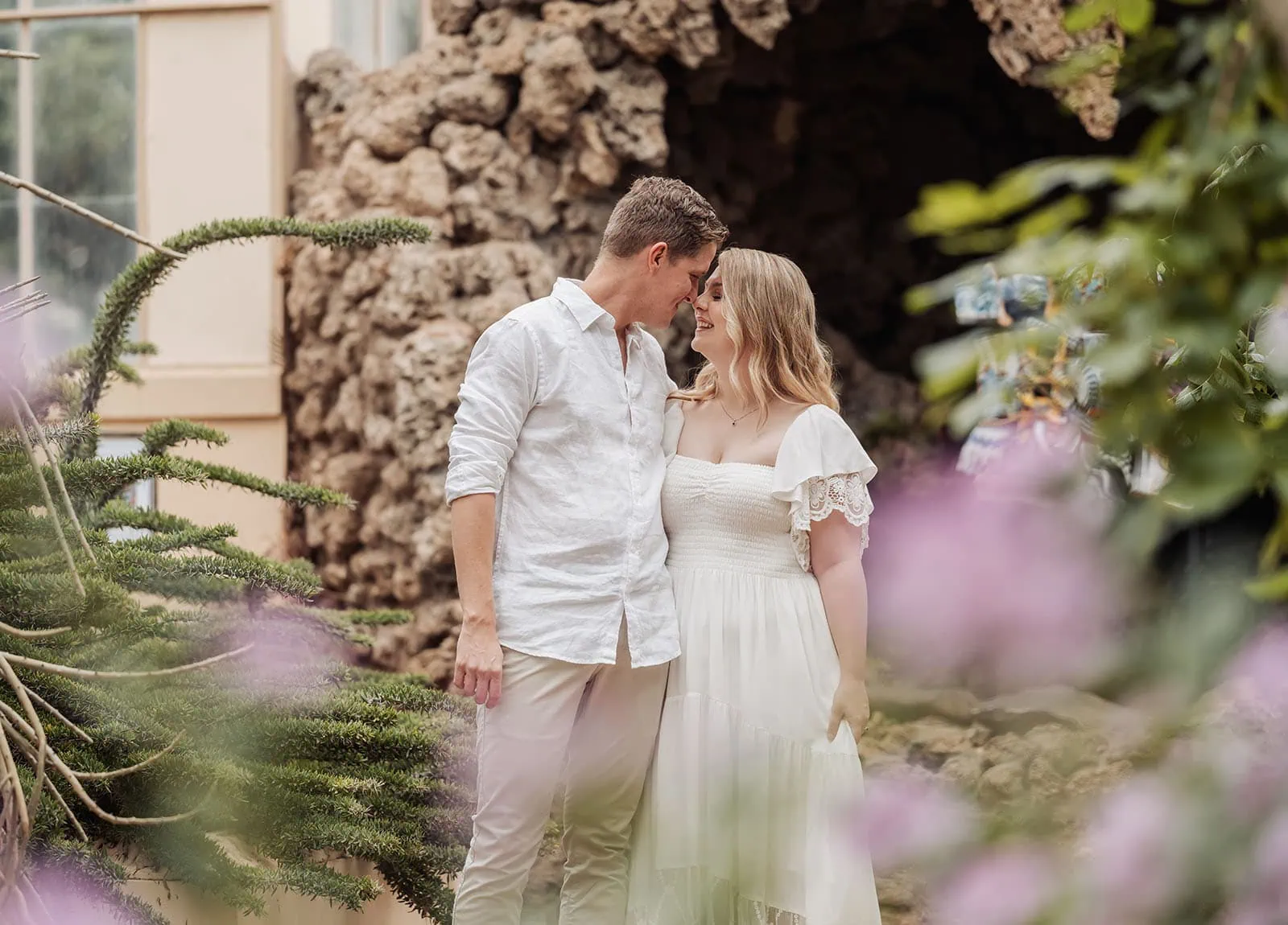botanic gardens wedding shoot - olivia & sean 11 Romantic couple in summer attire, embracing in a lush garden with rustic stone backdrop.
