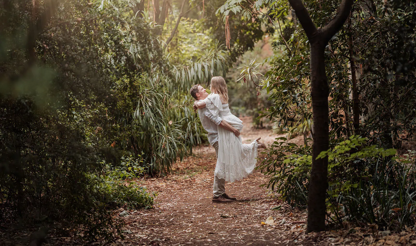 botanic gardens wedding shoot - olivia & sean 26 Romantic couple in white attire embracing in a sunlit, lush green forest.