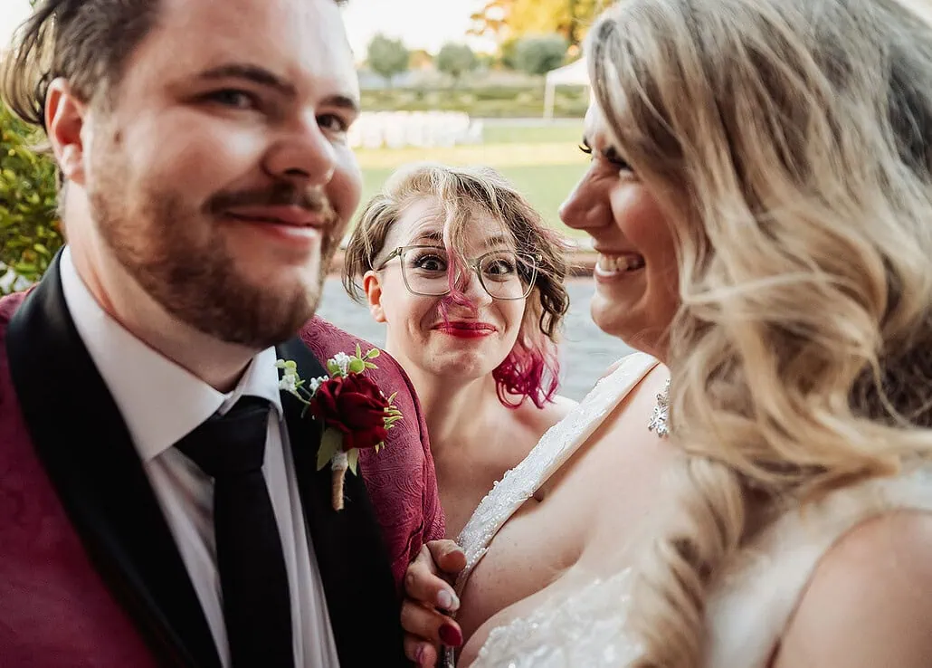 Bride and groom celebrate their wedding, joined by a playful photobomber in a garden setting.