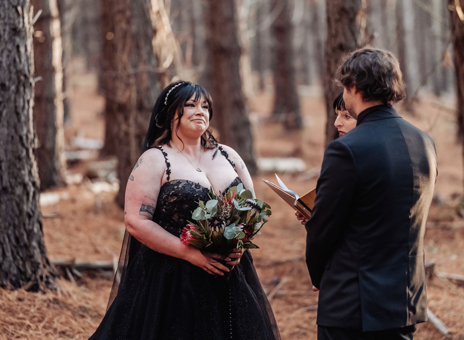 kuitpo forest metal themed wedding – erin & michael 2 Black wedding dress forest ceremony with bride, groom, and officiant exchanging vows.